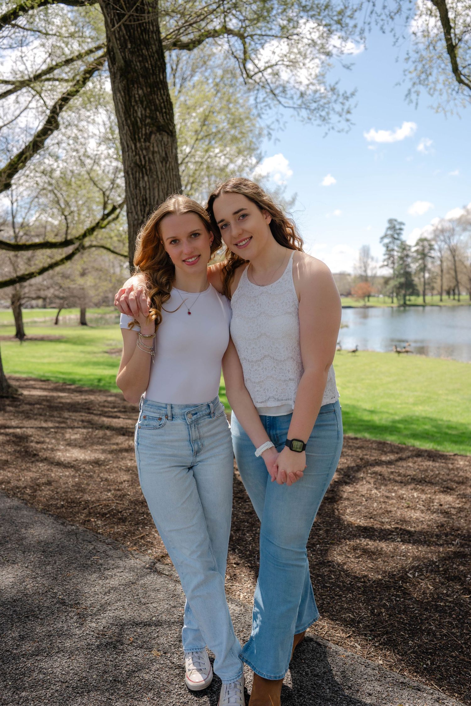 Two smiling young women pose together in a sunny park with a pond and green grass behind them.