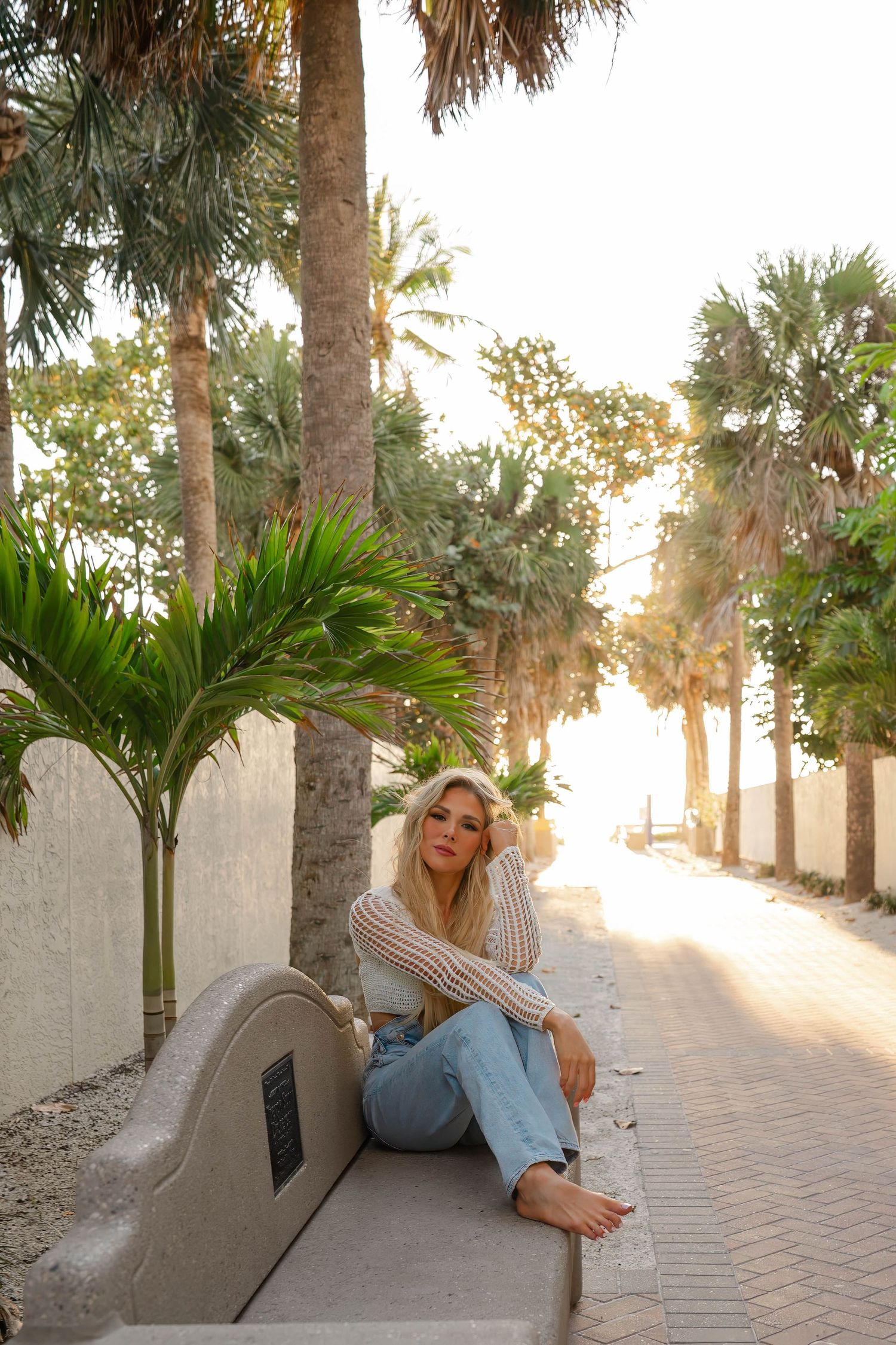 Blonde woman in white knit top and light jeans sitting barefoot on a bench along a palm tree-lined path.
