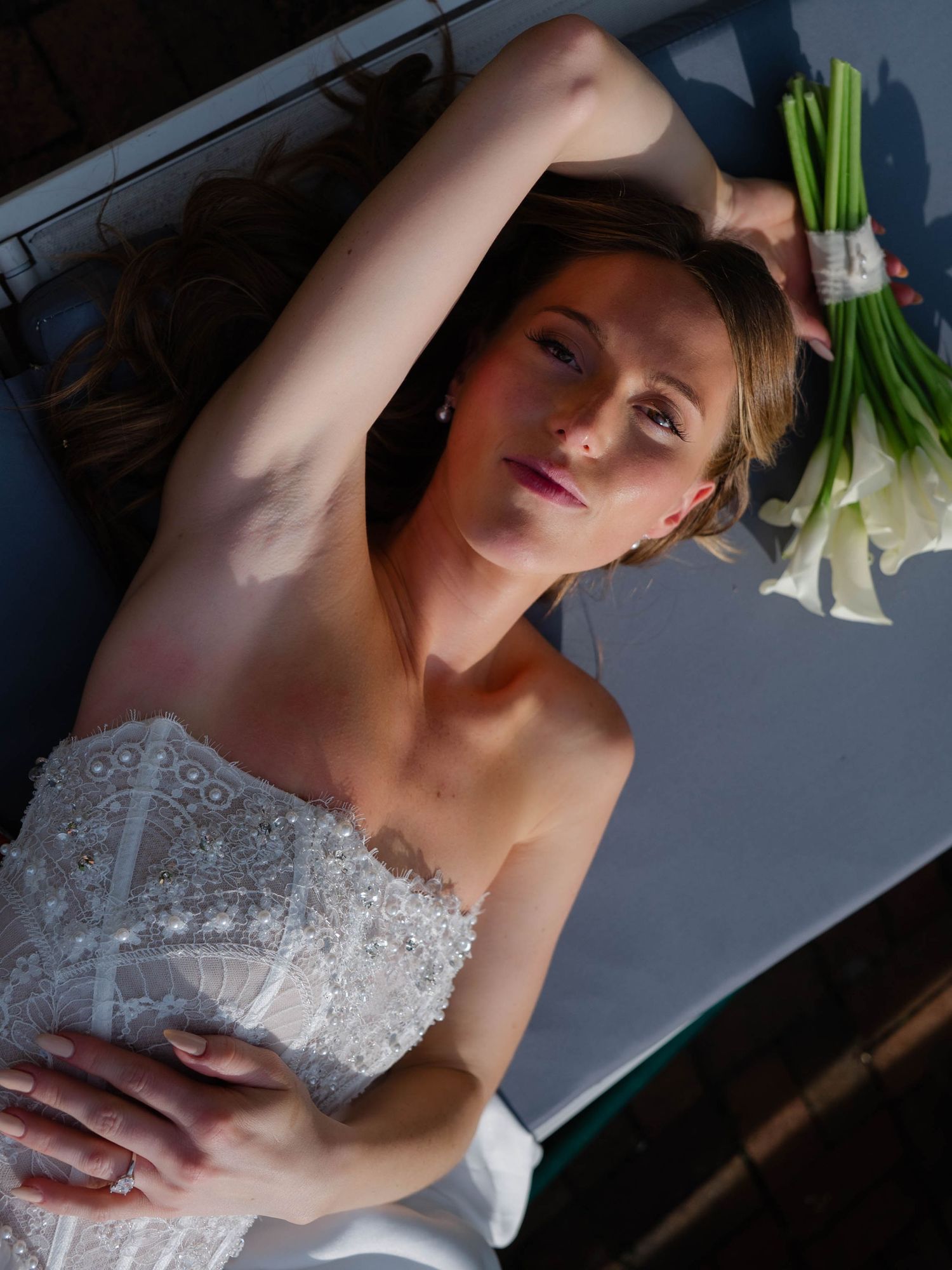 Close-up of bride lying back holding calla lily bouquet in dramatic sunlight wearing lace gown.