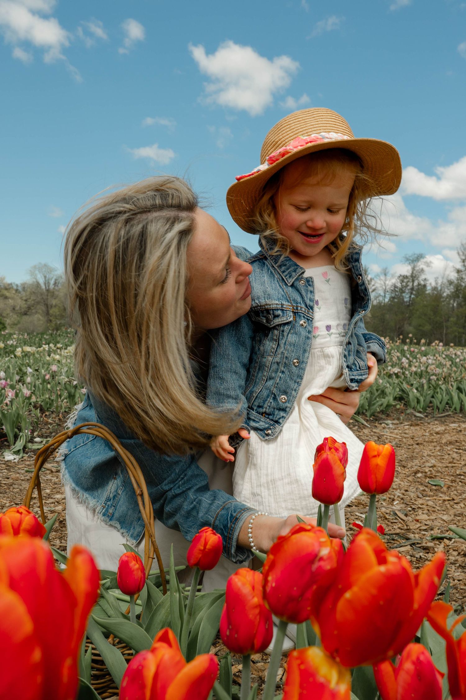 Mother holding young girl in straw hat among vibrant red tulips in spring flower field.