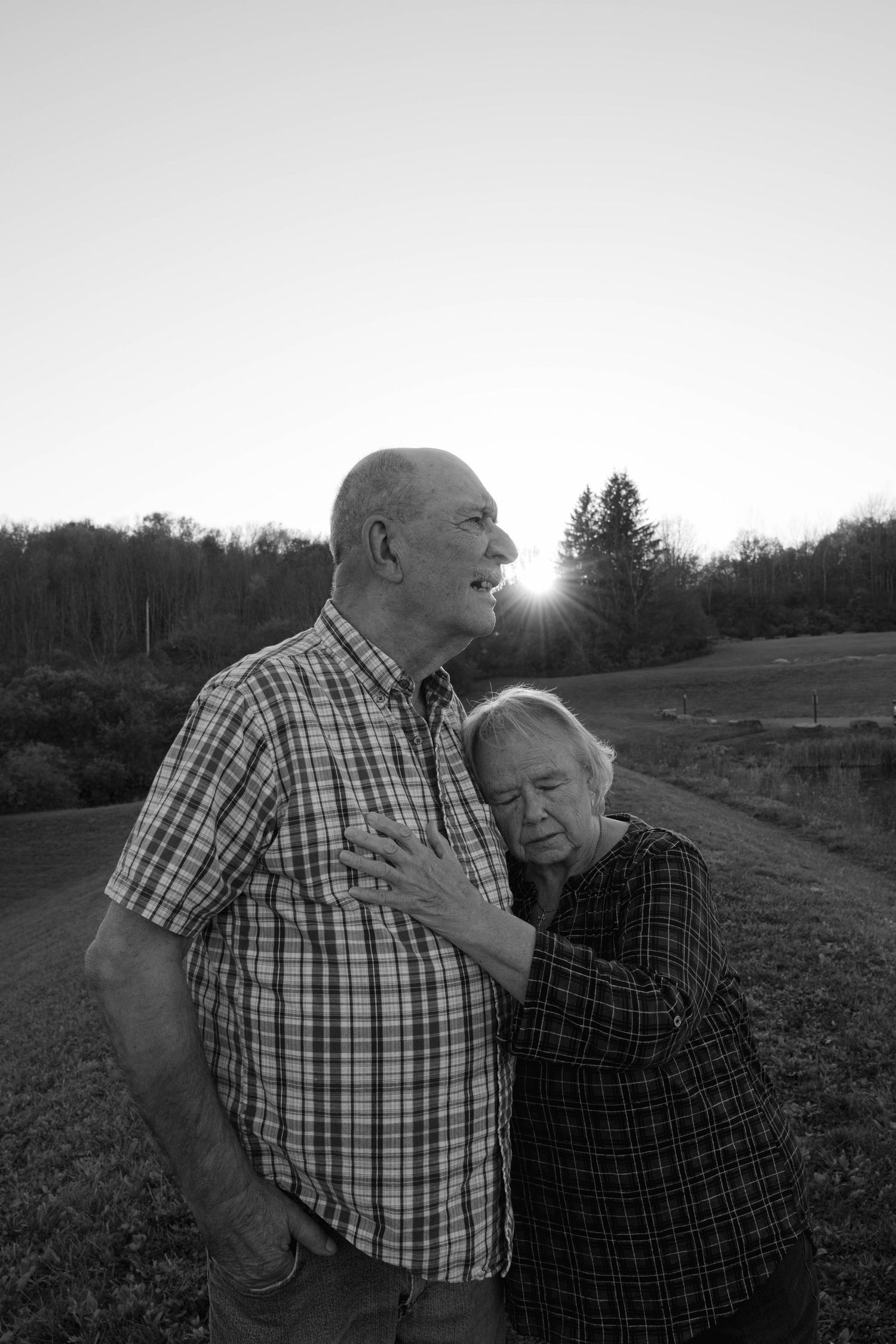 Black and white portrait of an elderly couple embracing at sunset with rolling hills in background.