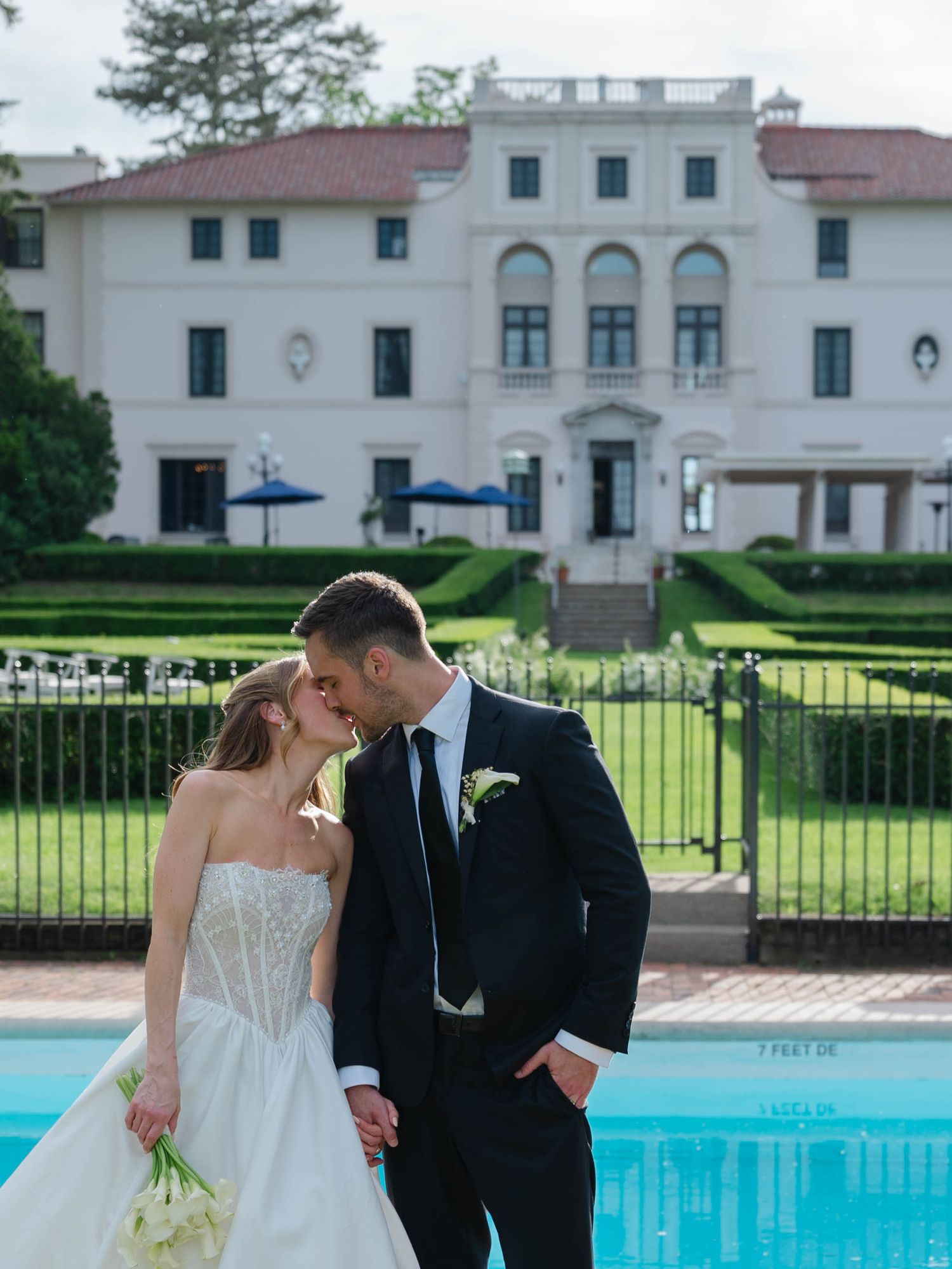 Newlyweds share a romantic kiss poolside in front of an elegant white estate with lush green hedges.