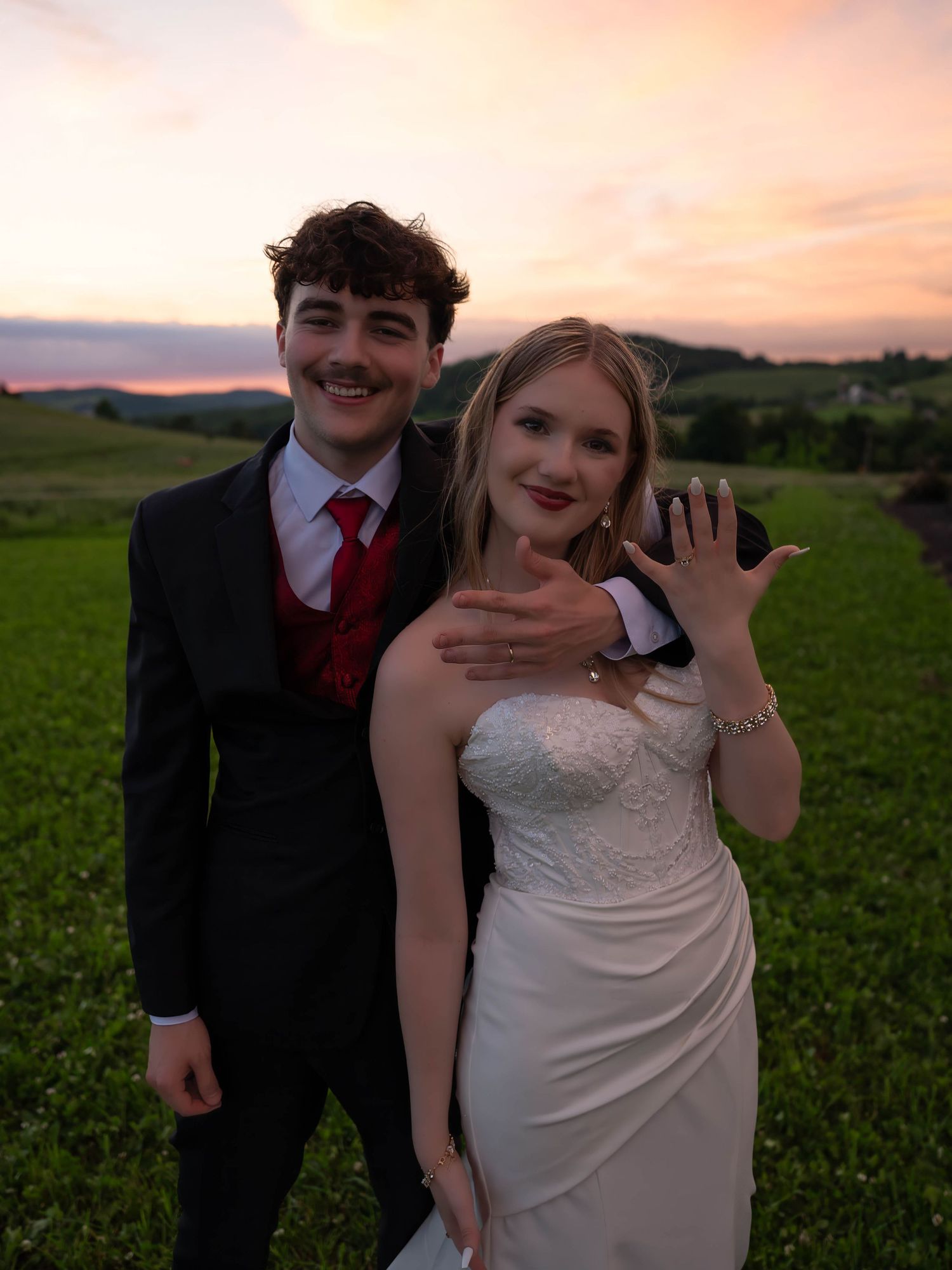 Newlywed couple posing at sunset in green hills, groom in black tuxedo with red tie, bride in white gown showing wedding rings.