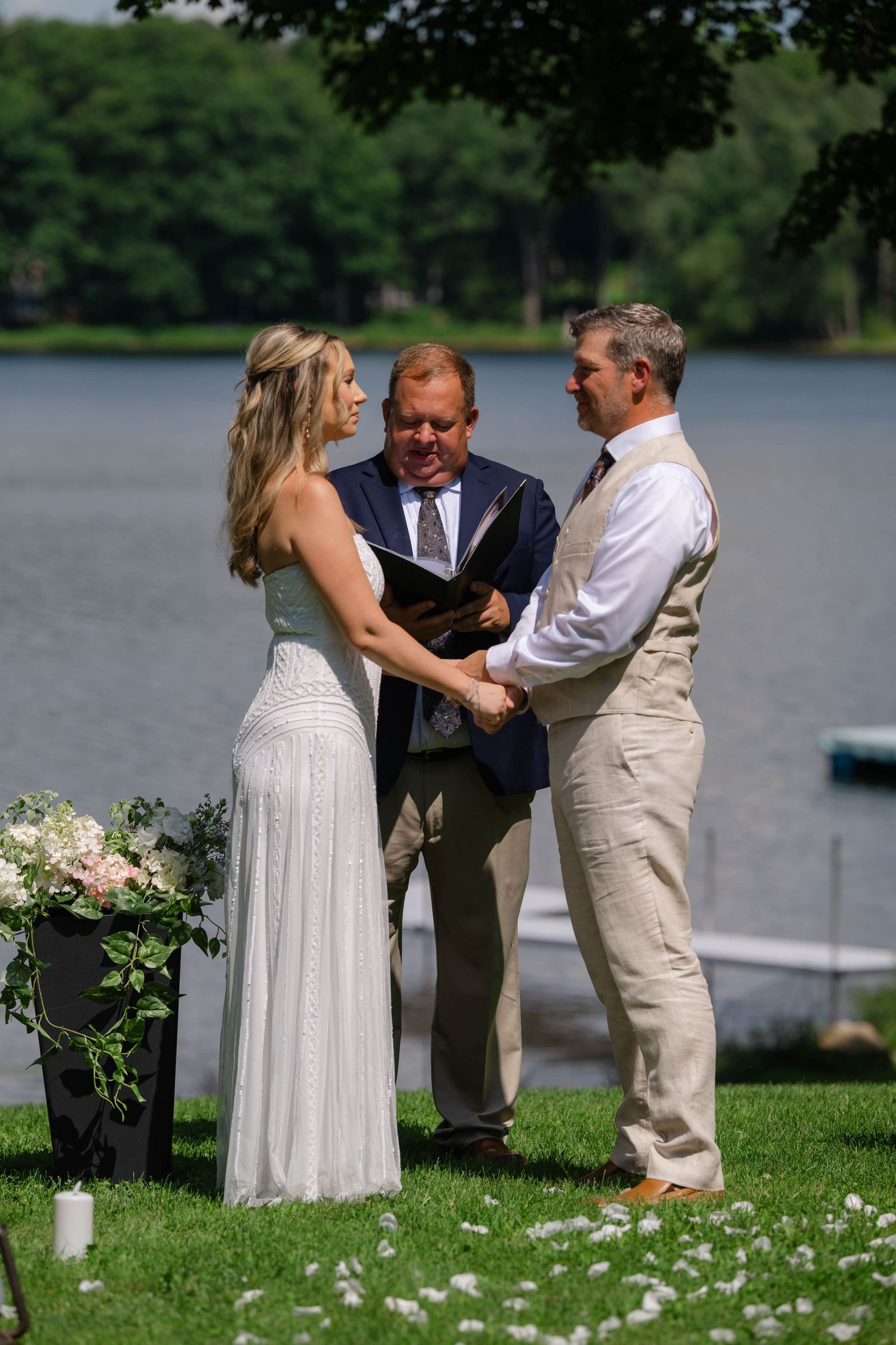 Outdoor lakeside wedding ceremony with bride and groom holding hands before officiant on sunny day.