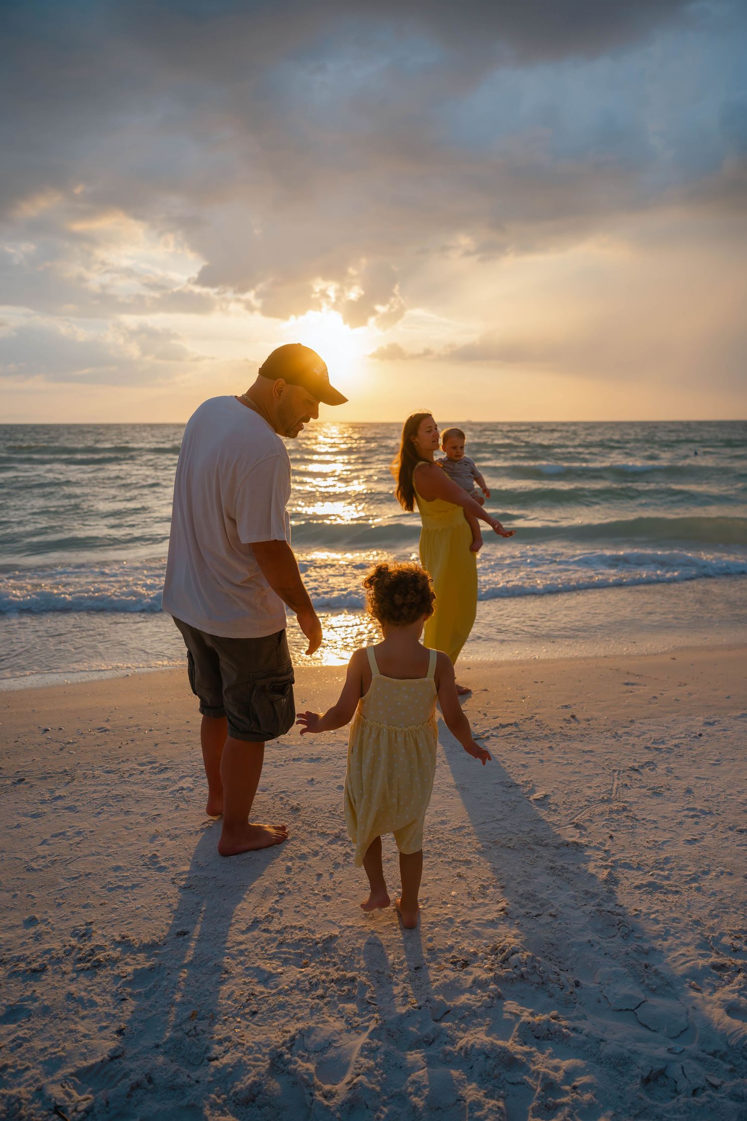 Family walking on a white sand beach at golden sunset, parents with two young children near ocean waves.