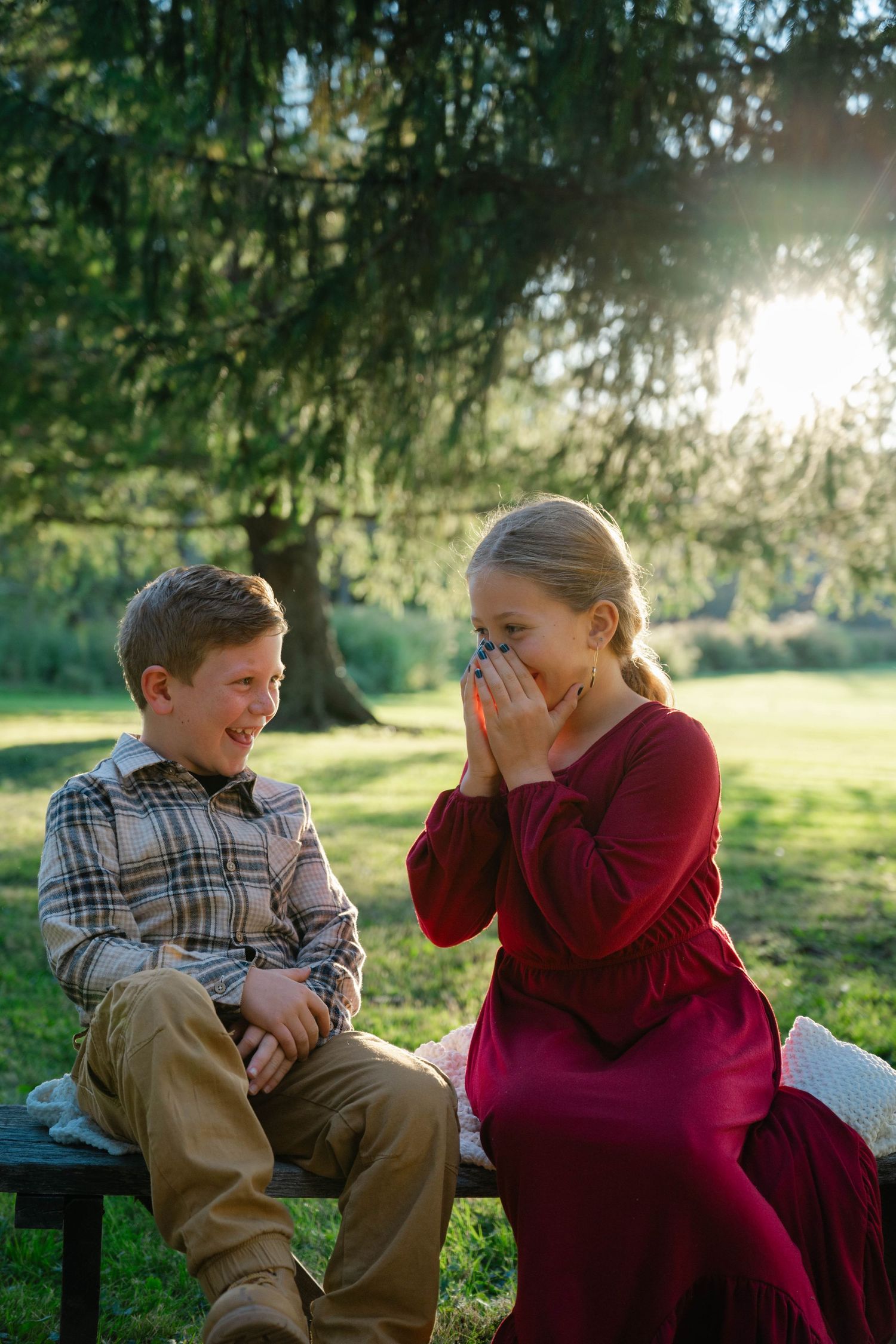 Two children laughing together on a bench in a sunlit park, boy in plaid shirt and girl in red dress.