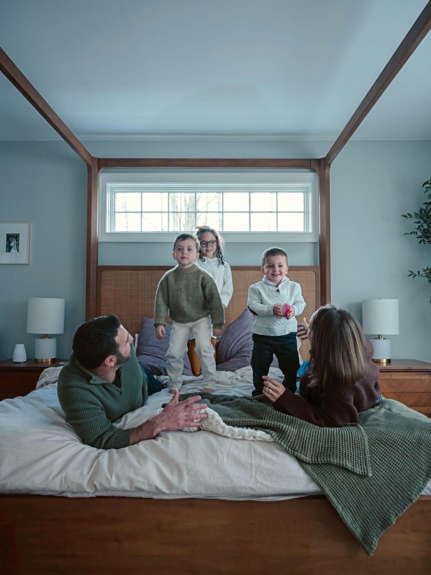 Four children stand and smile on a bed while their parents relax in front of them.