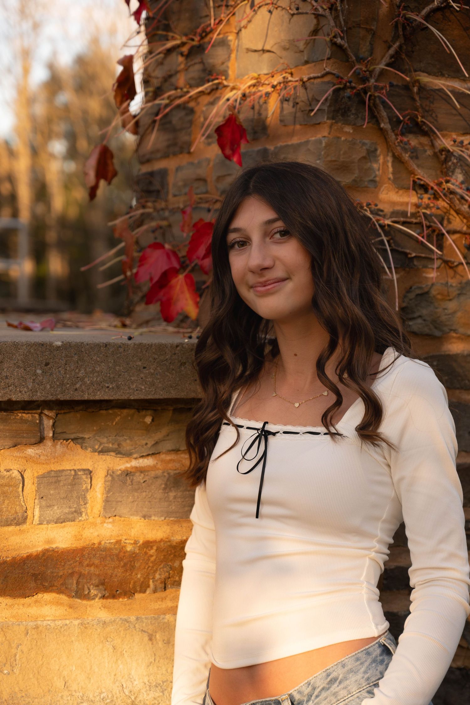 Teen girl with long wavy hair smiling beside stone wall with red autumn leaves in warm sunlight.