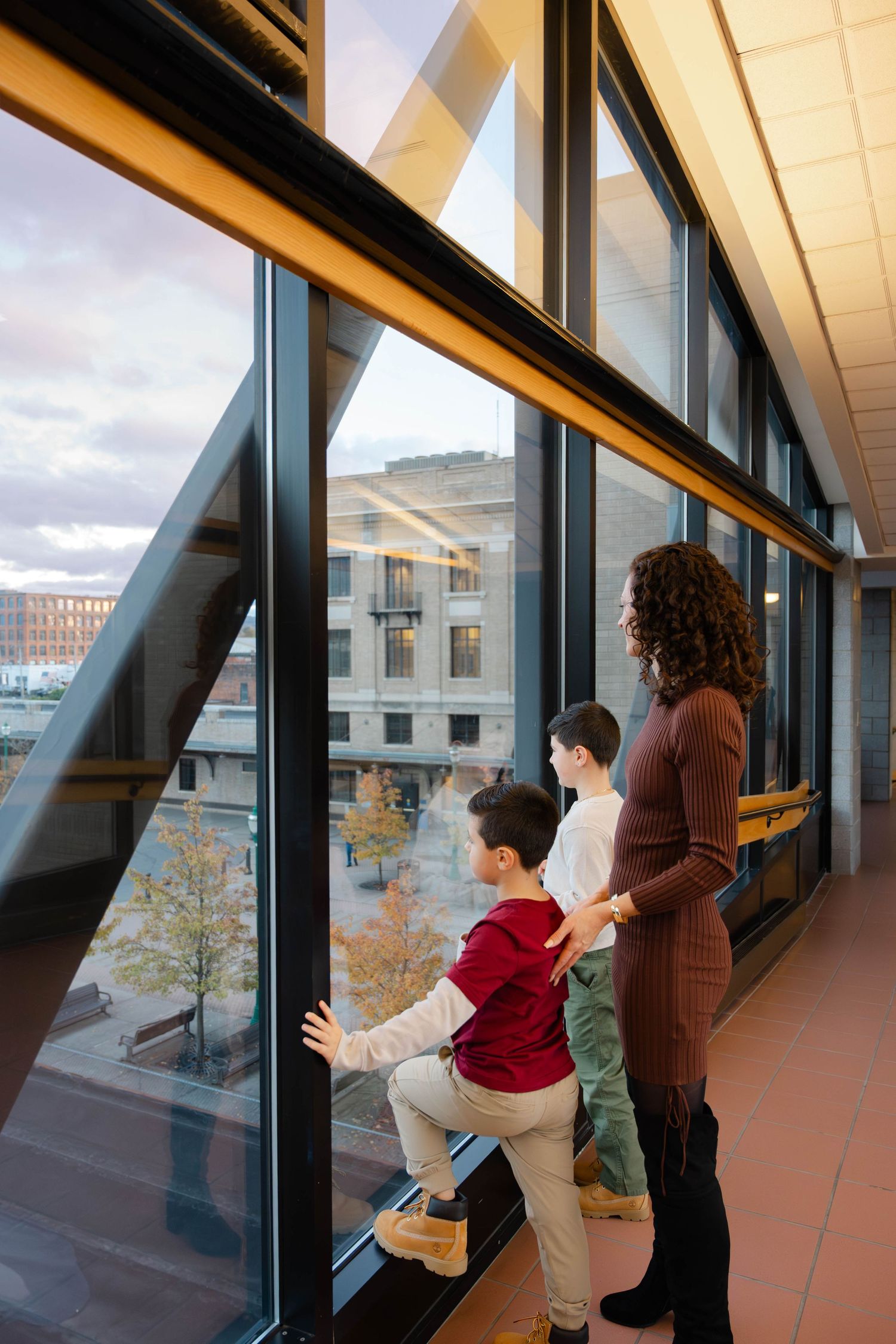 Pregnant mother and two sons look out large windows at city view in modern building hallway.