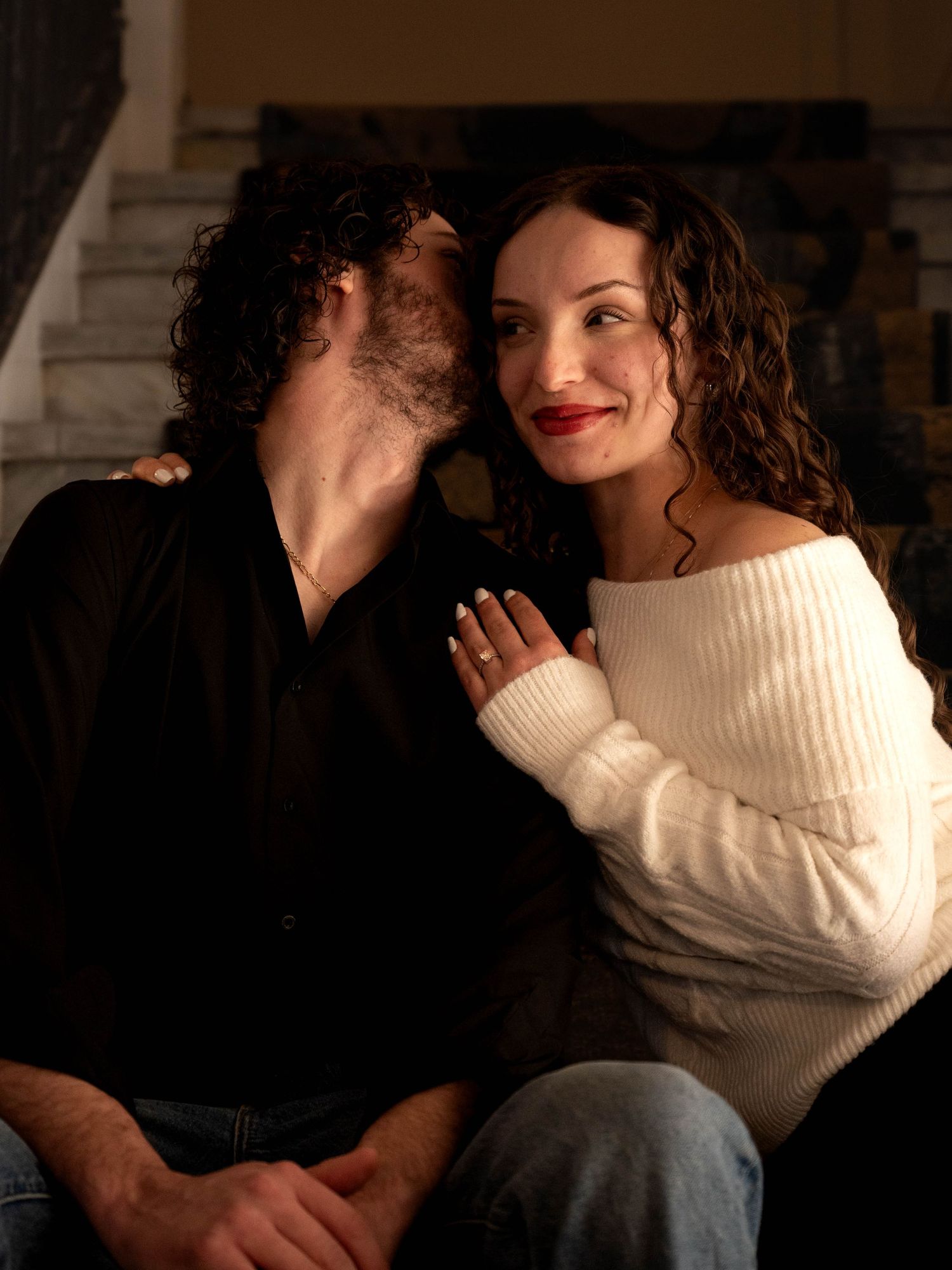 A smiling woman with red lips and curly hair leans into her partner near a stone fireplace.