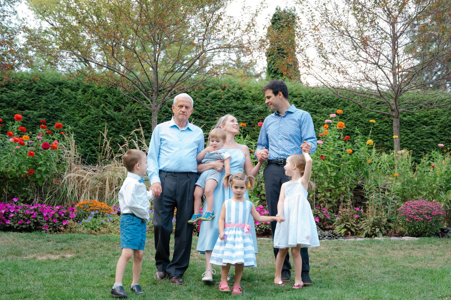 Multi-generational family portrait with grandparents, parents, and young children posing in a lush garden with colorful flowers.