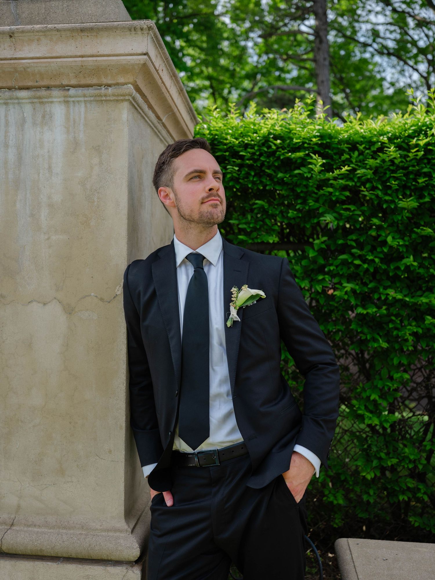 Dapper groom in black suit with boutonniere leans against a stone column surrounded by green hedges.