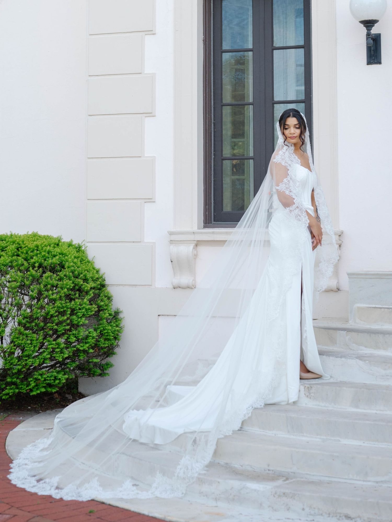 Elegant bride in fitted lace wedding gown with long cathedral veil posing on stone steps outside a white mansion.