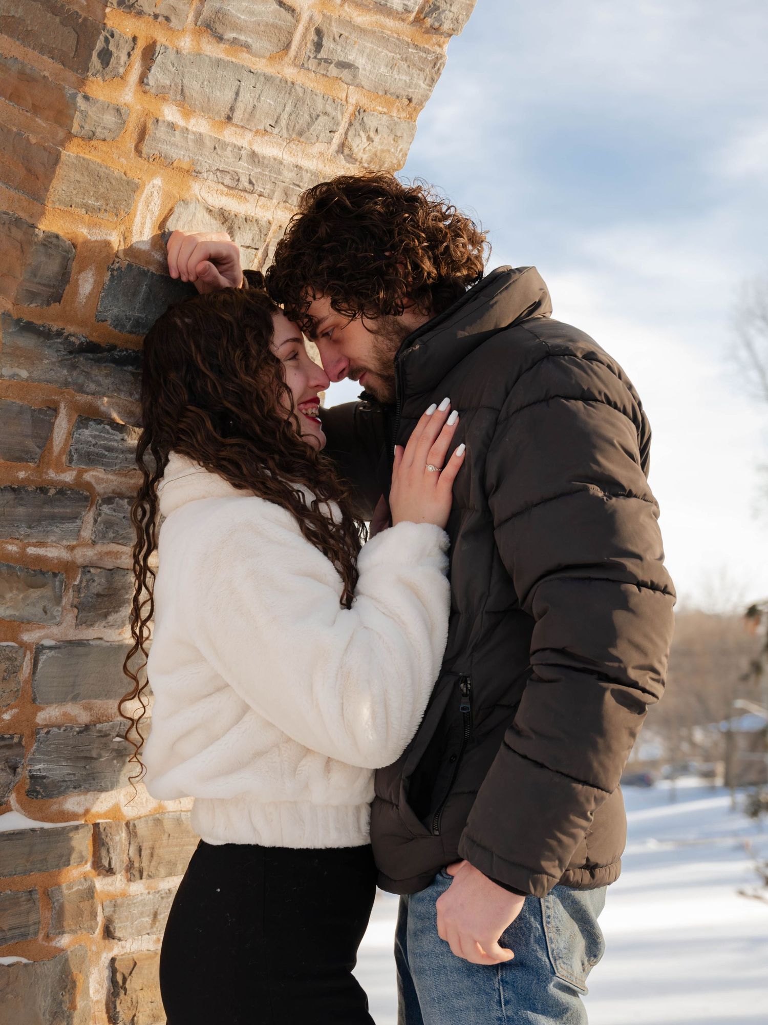 Couple nearly kissing against a stone wall in a snowy winter outdoor setting.