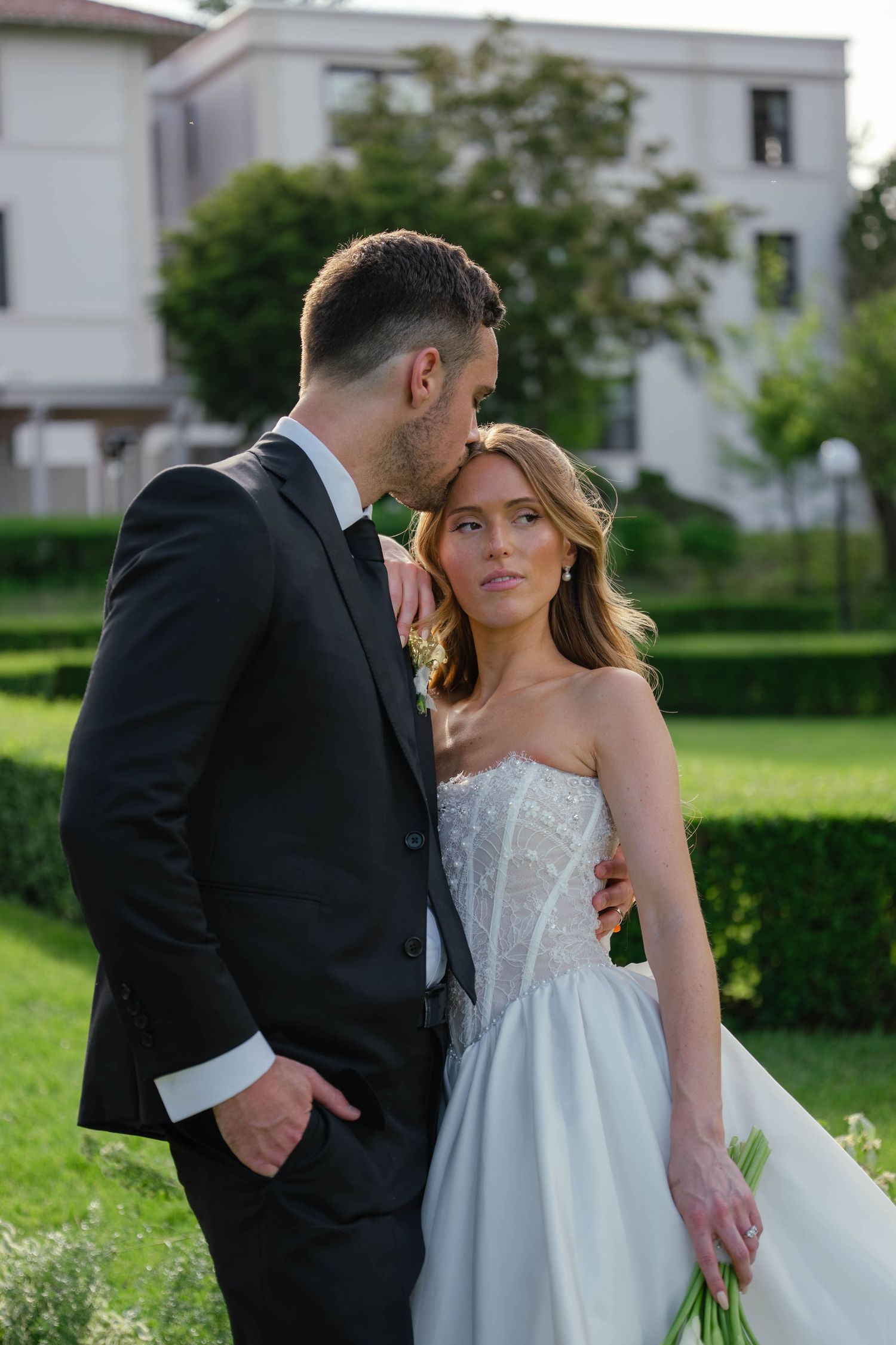 Groom in black tuxedo embracing bride in strapless corset ball gown in lush green garden.