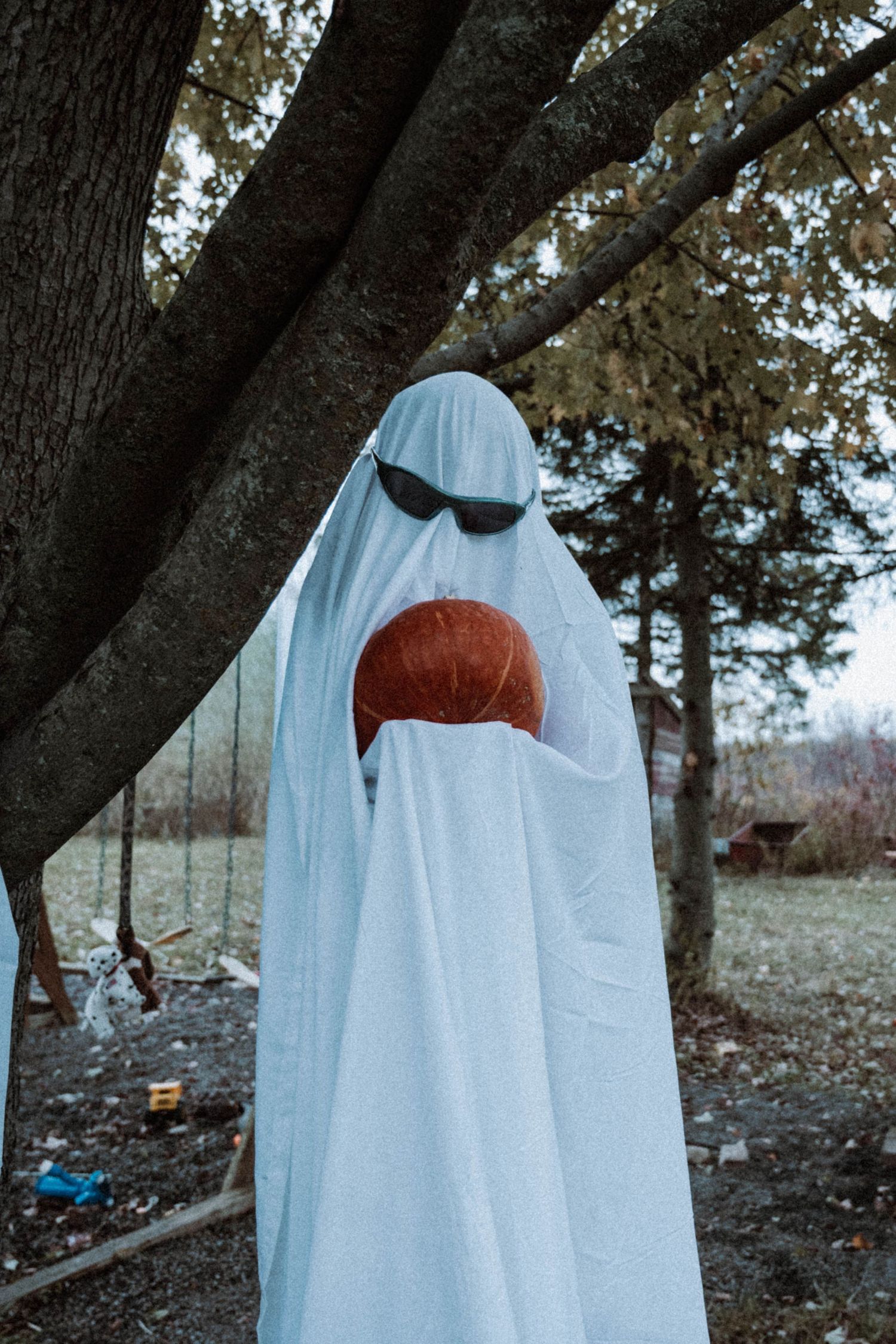 Person in white ghost costume with sunglasses holding a pumpkin outdoors near a tree during Halloween.