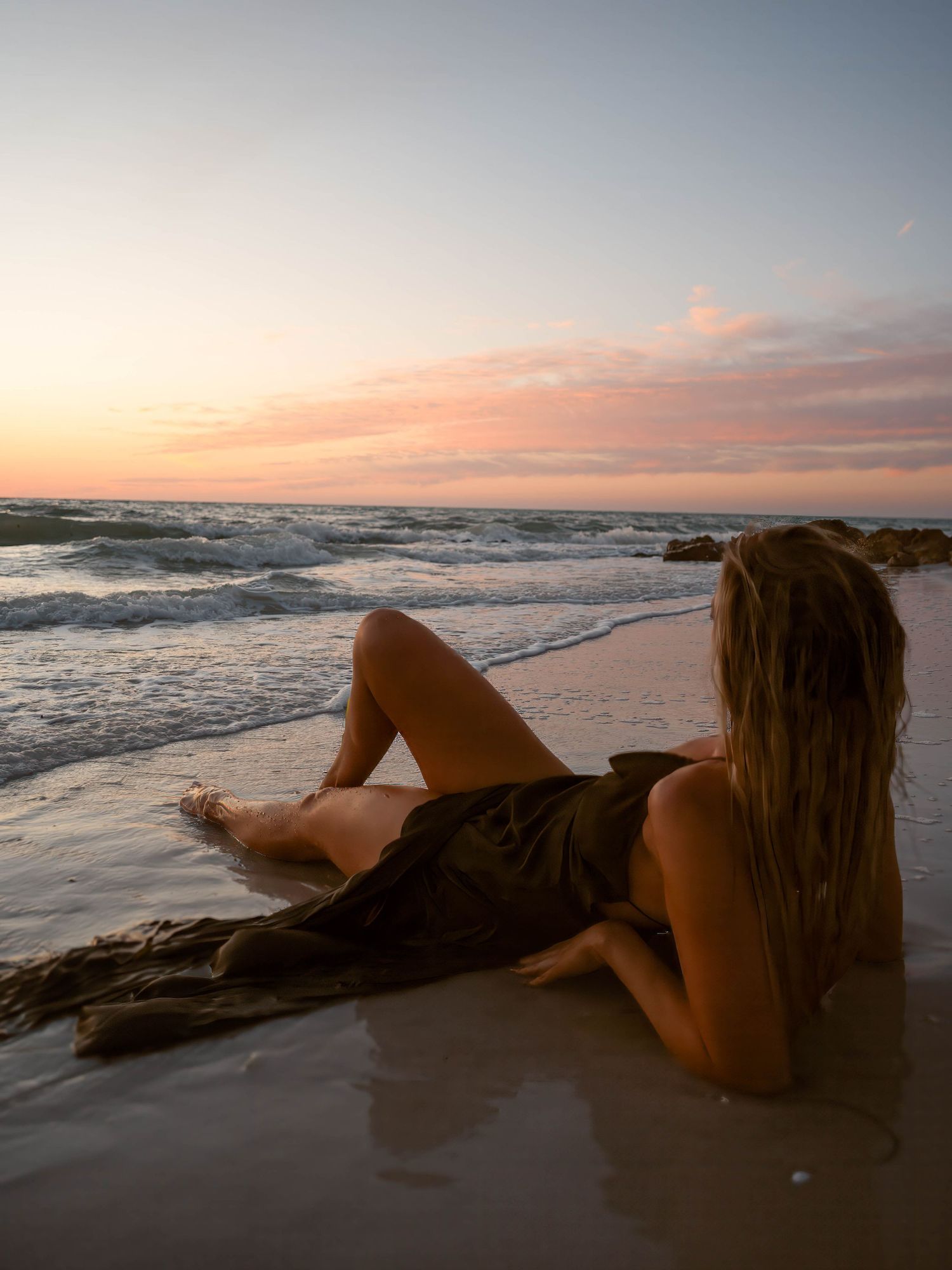 Woman in olive wrap dress lounging on sandy beach at sunset with ocean waves in background.