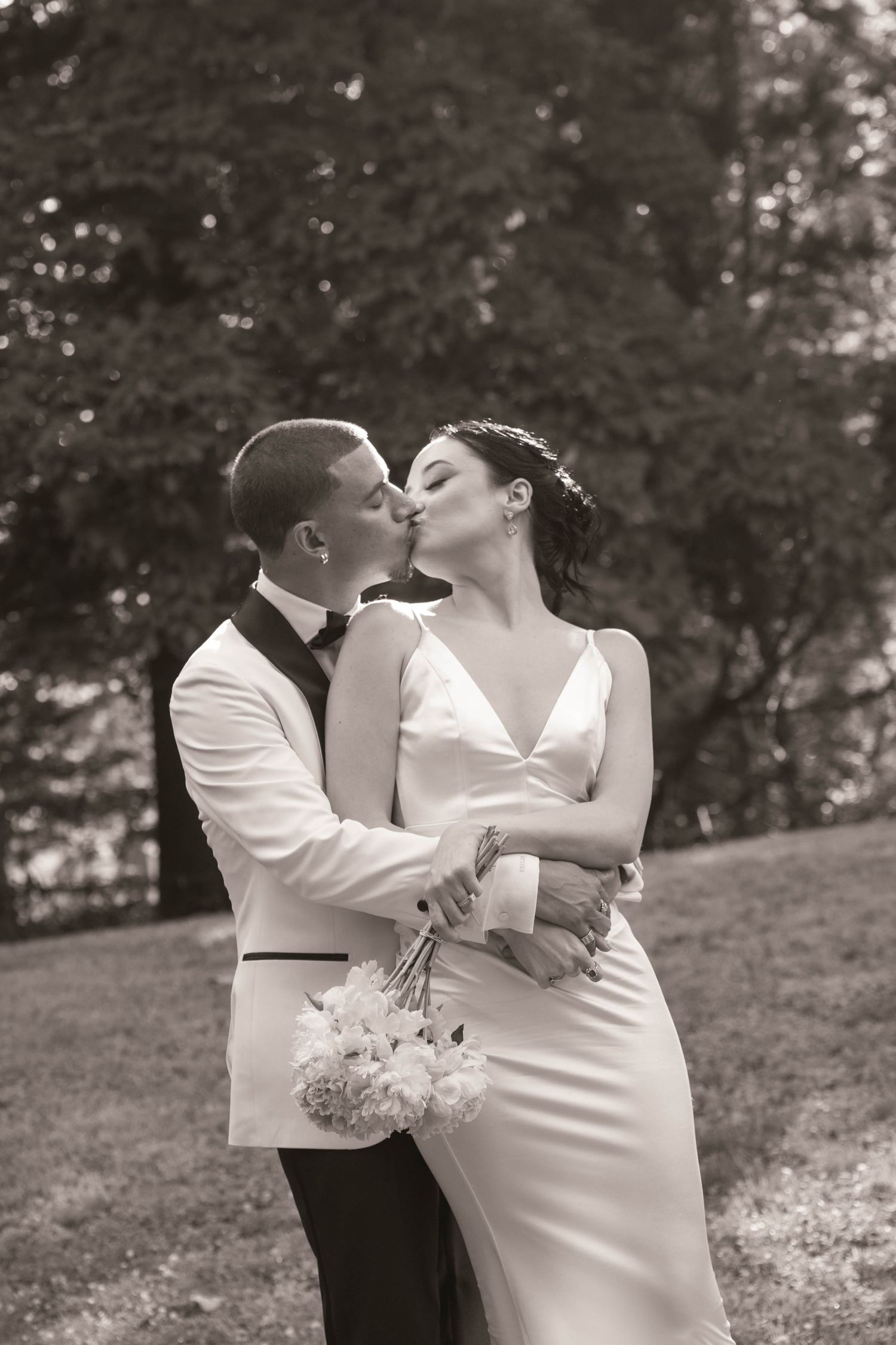 Black and white photo of a bride and groom kissing outdoors, she holds a bouquet in a sleek white gown, he wears a white tuxedo.