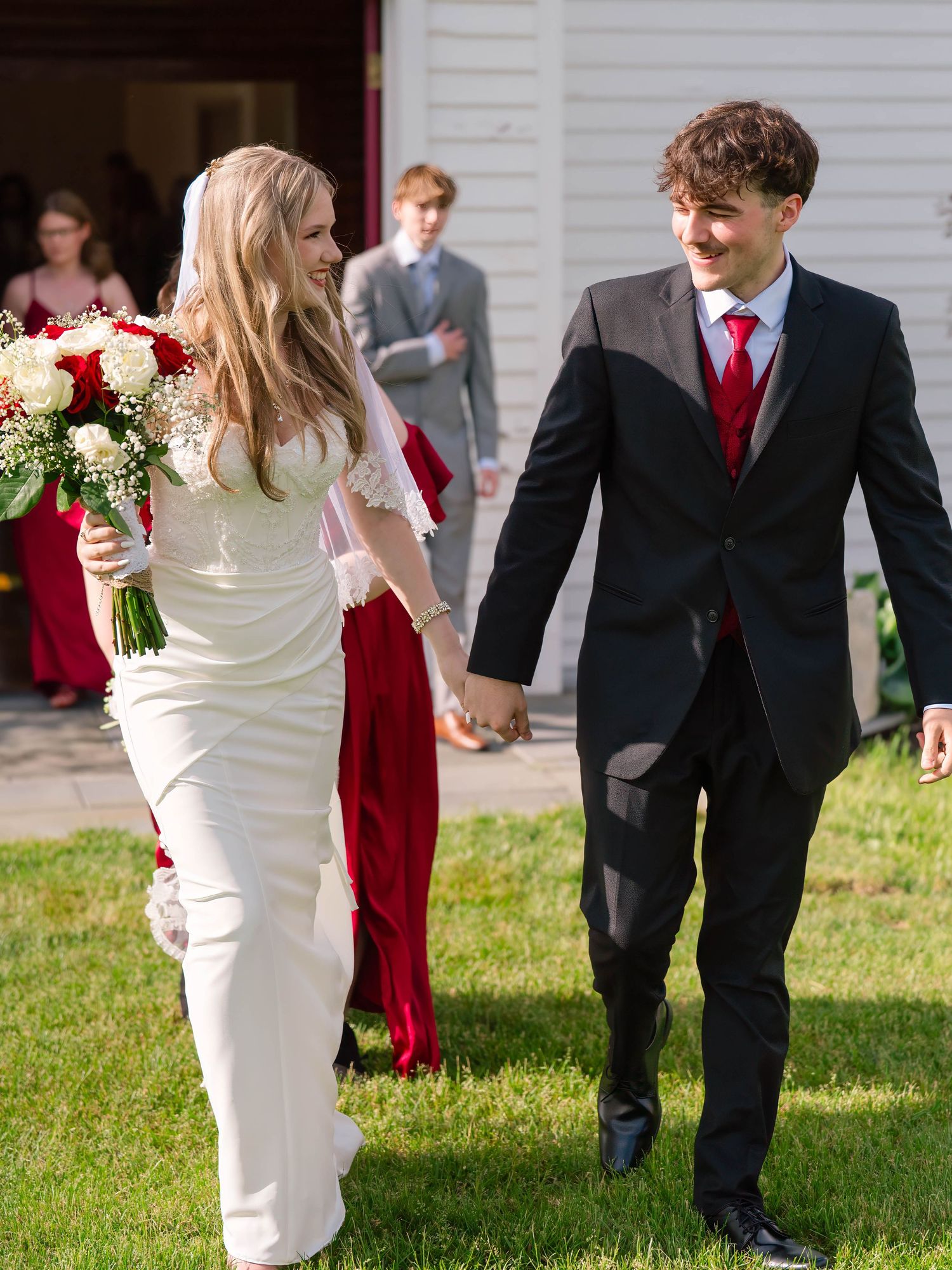 Bride in white dress and groom in black suit with red tie hold hands walking on grass at outdoor wedding.
