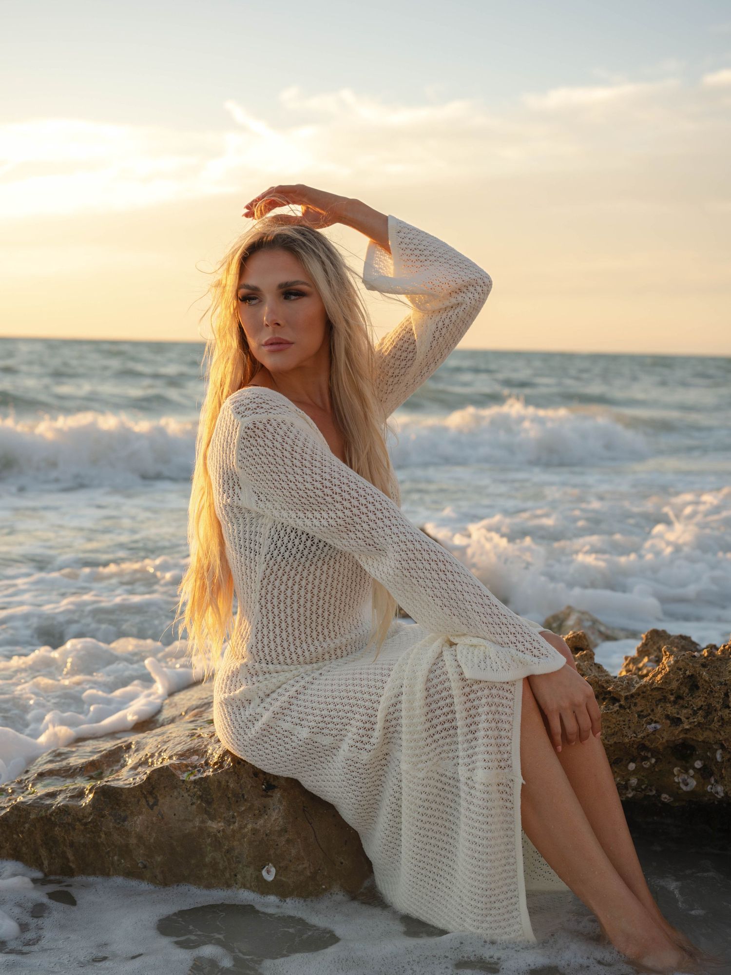 Blonde woman in white crochet dress sitting on rocks at sunset beach with ocean waves in background.