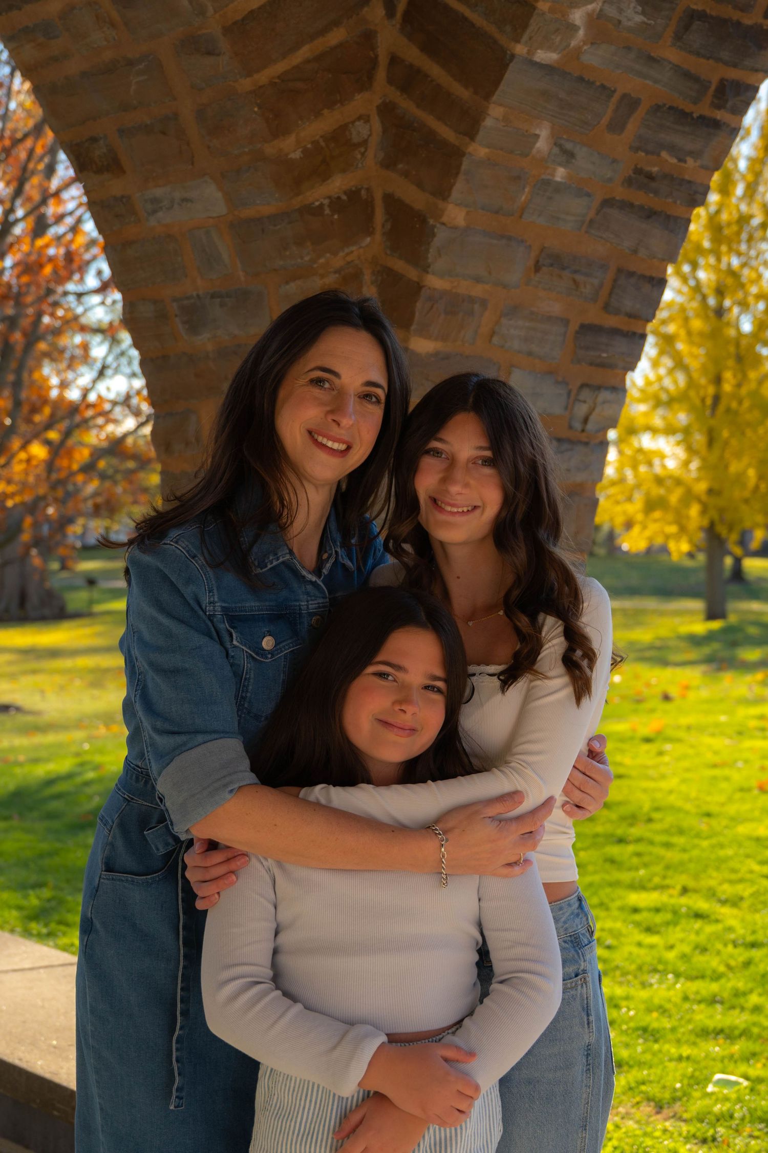 Three women smile and embrace under a stone arch in a sunny autumn park with golden foliage.