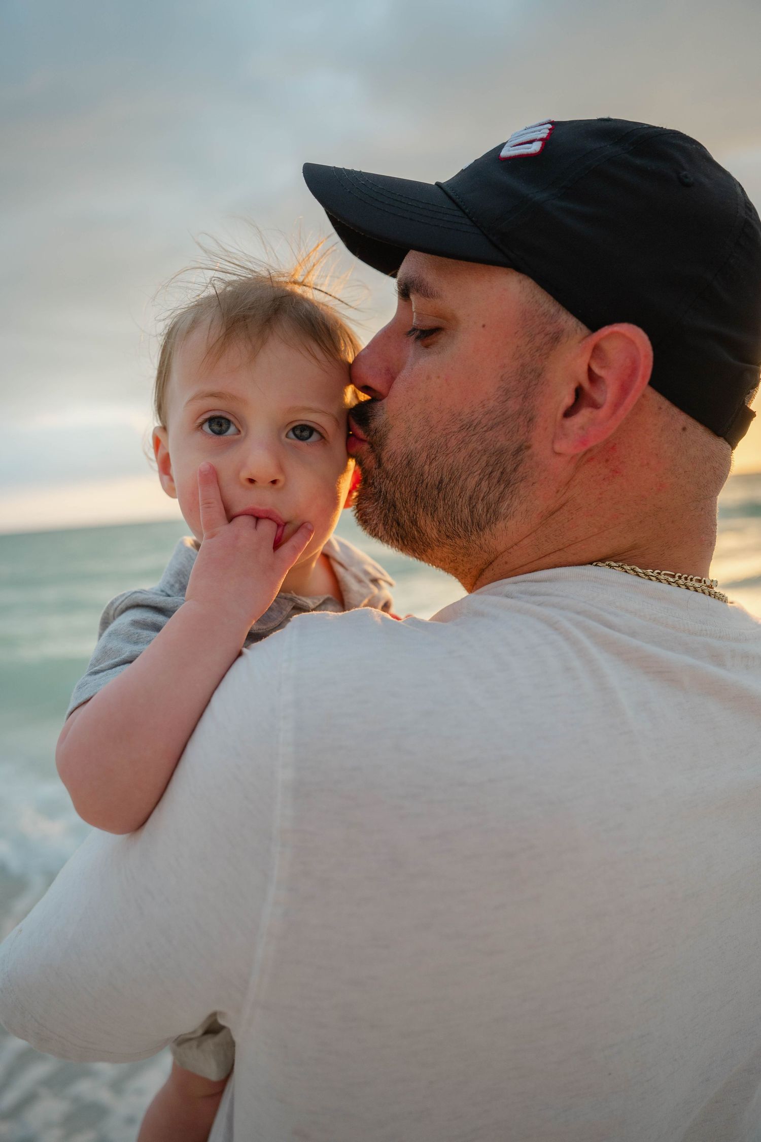 Father kissing young toddler on cheek at sunset beach, child with finger in mouth looking at camera.