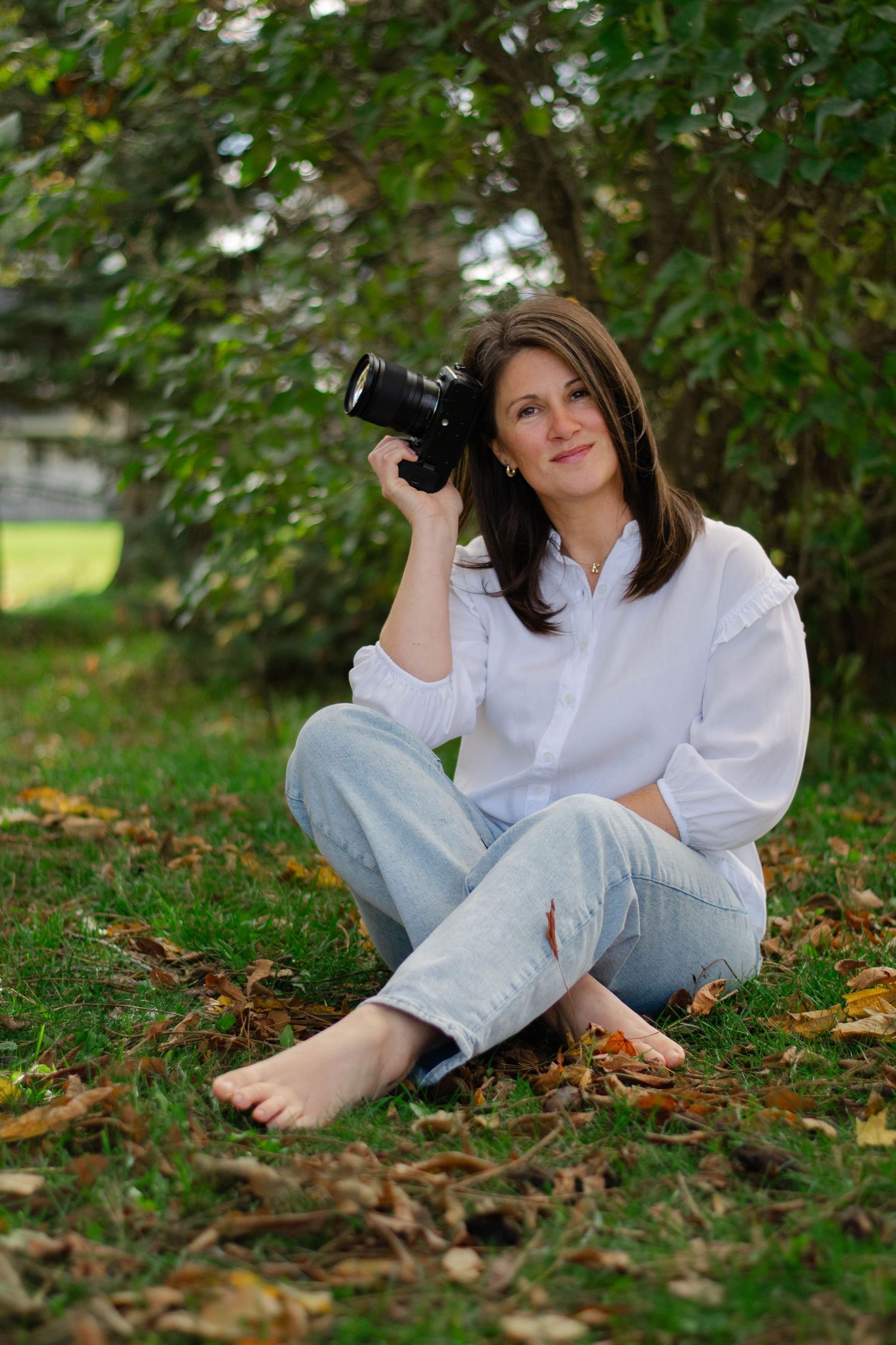 Female photographer sitting barefoot on grass holding camera, smiling among autumn leaves under trees.