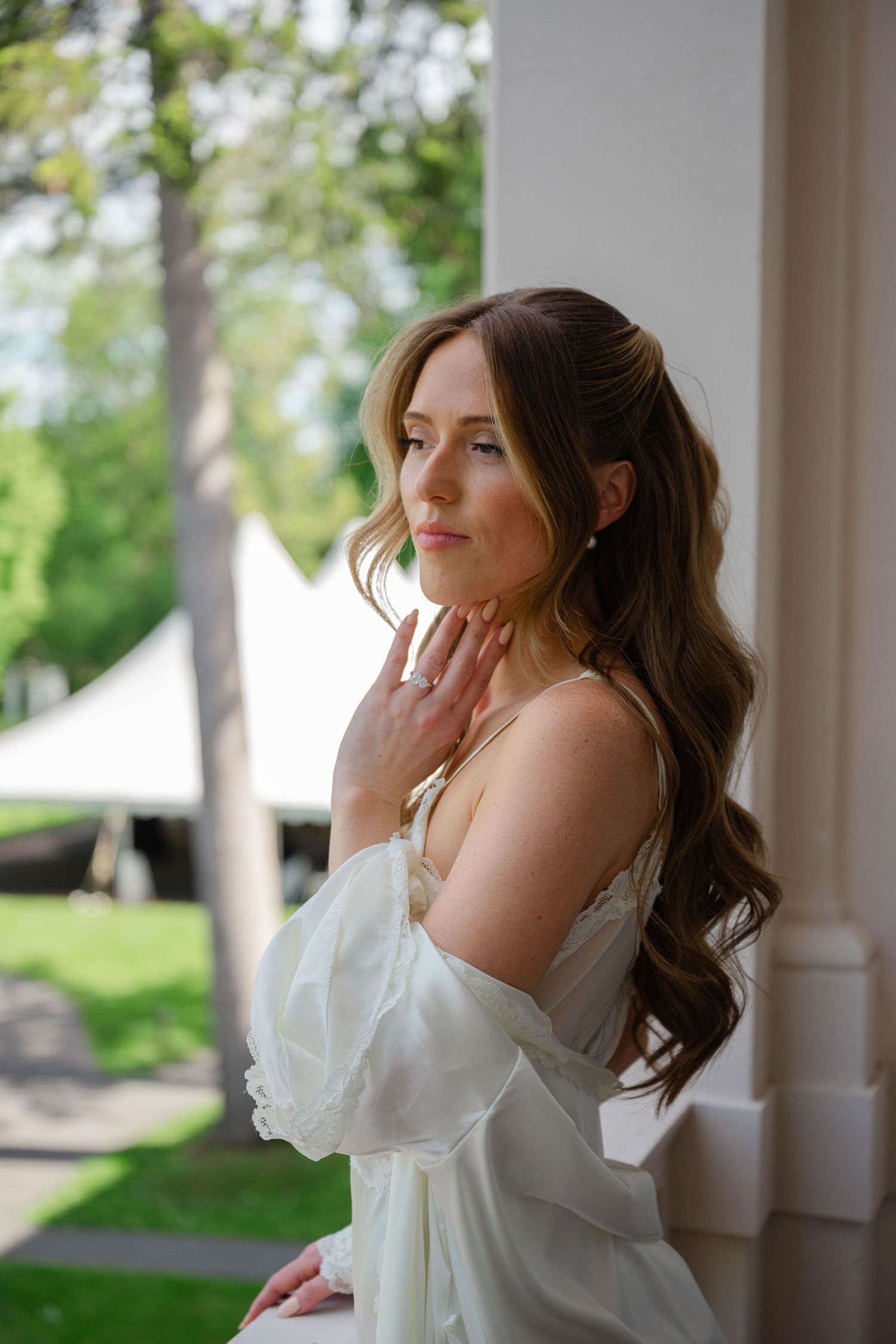 Beautiful bride in white off-shoulder gown touches her neck while posing near white column.