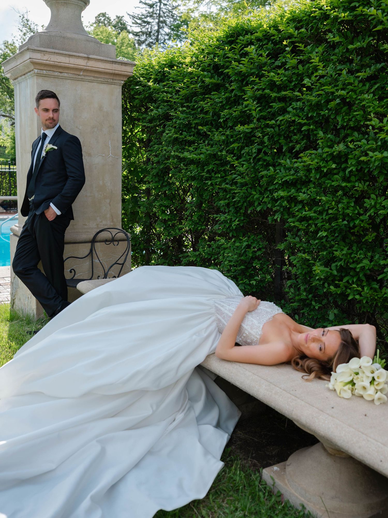 Bride in full white gown reclines on stone bench while groom stands by a pool column in lush garden.