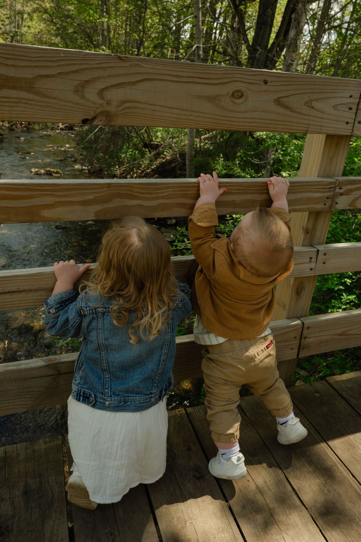 Two young children in denim and tan outfits peering through wooden bridge railing at stream below.