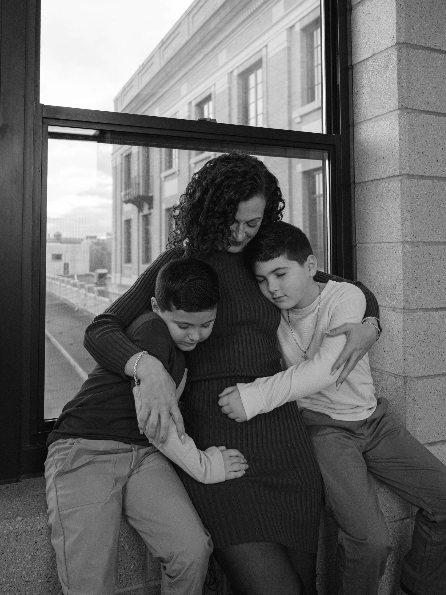 Black and white photo of two boys hugging their pregnant mother by a large window.