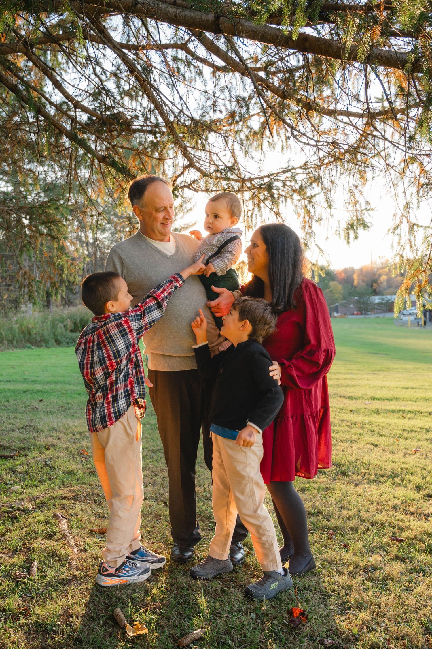 Happy family of five with baby stands under pine tree branches in golden autumn sunlight.