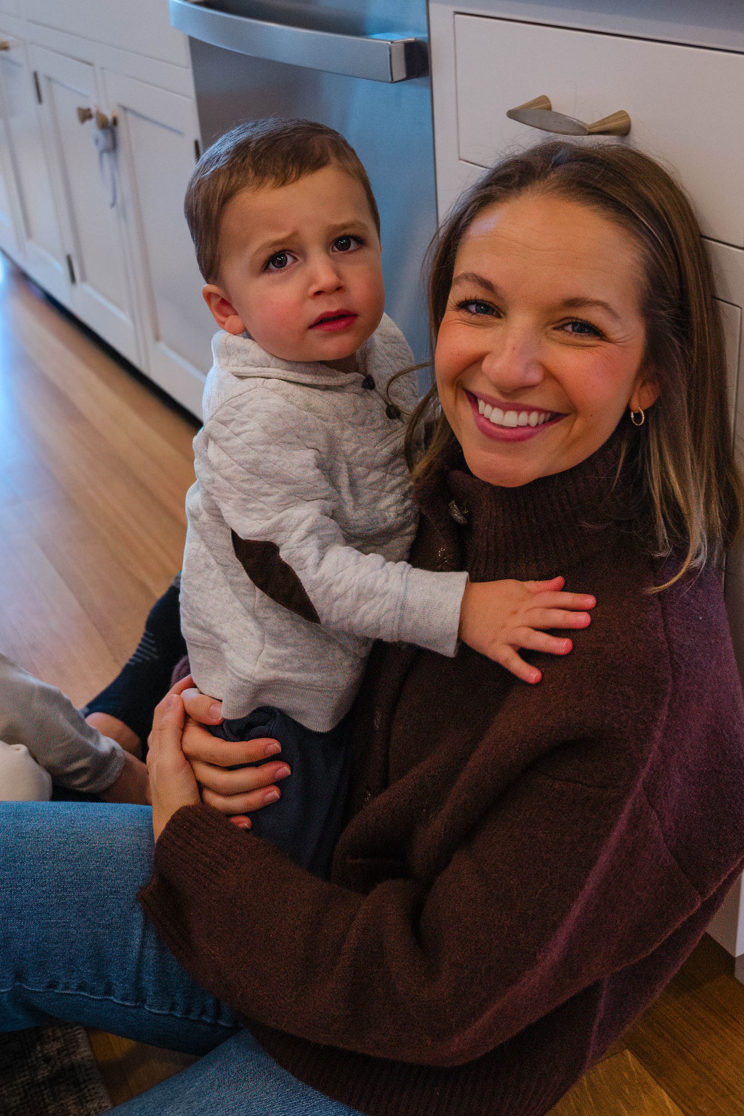 Smiling mother holds young toddler boy while sitting on kitchen floor in warm home setting.