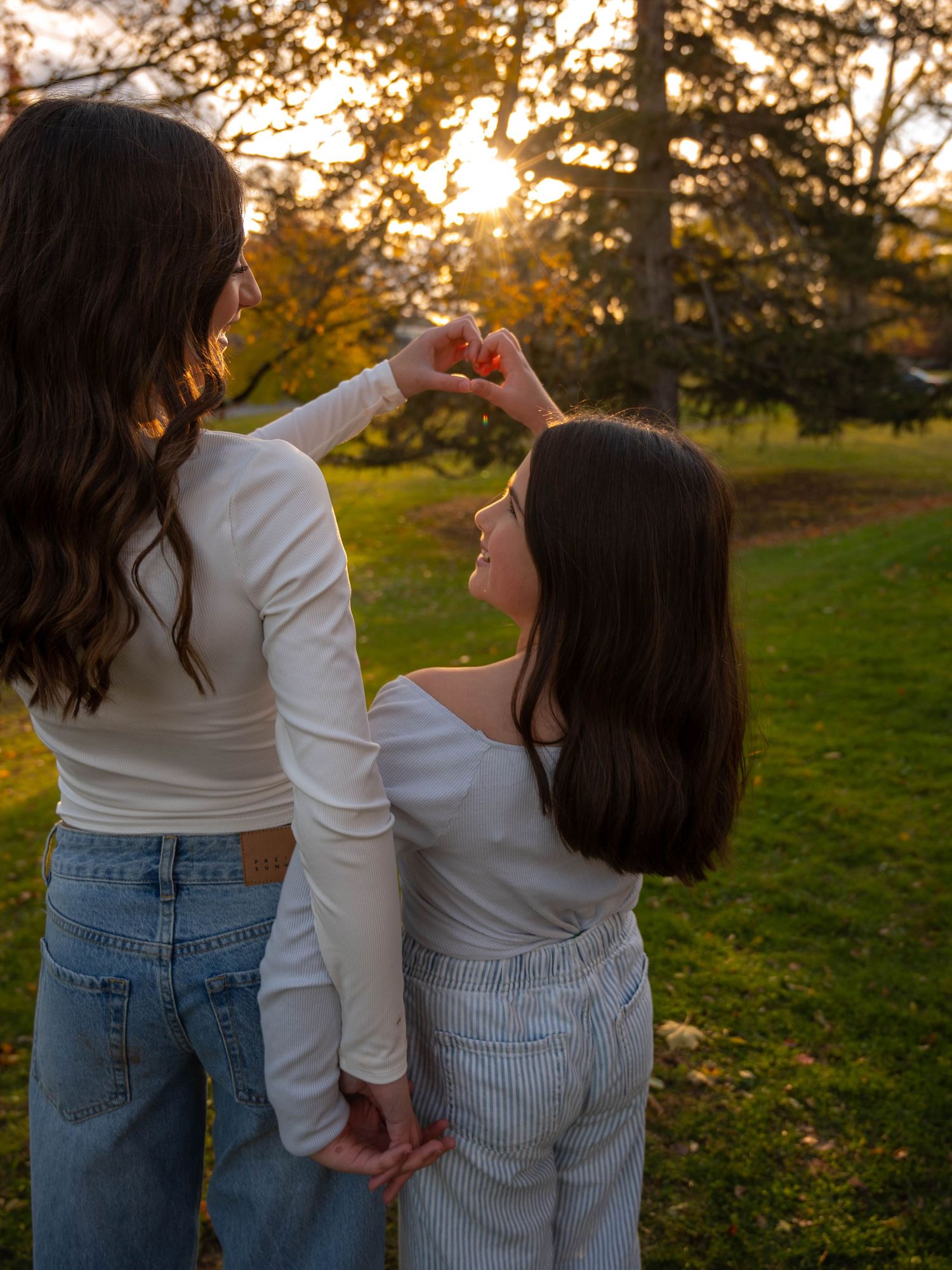 Two girls in white tops make a heart shape with their hands at golden sunset in a park.