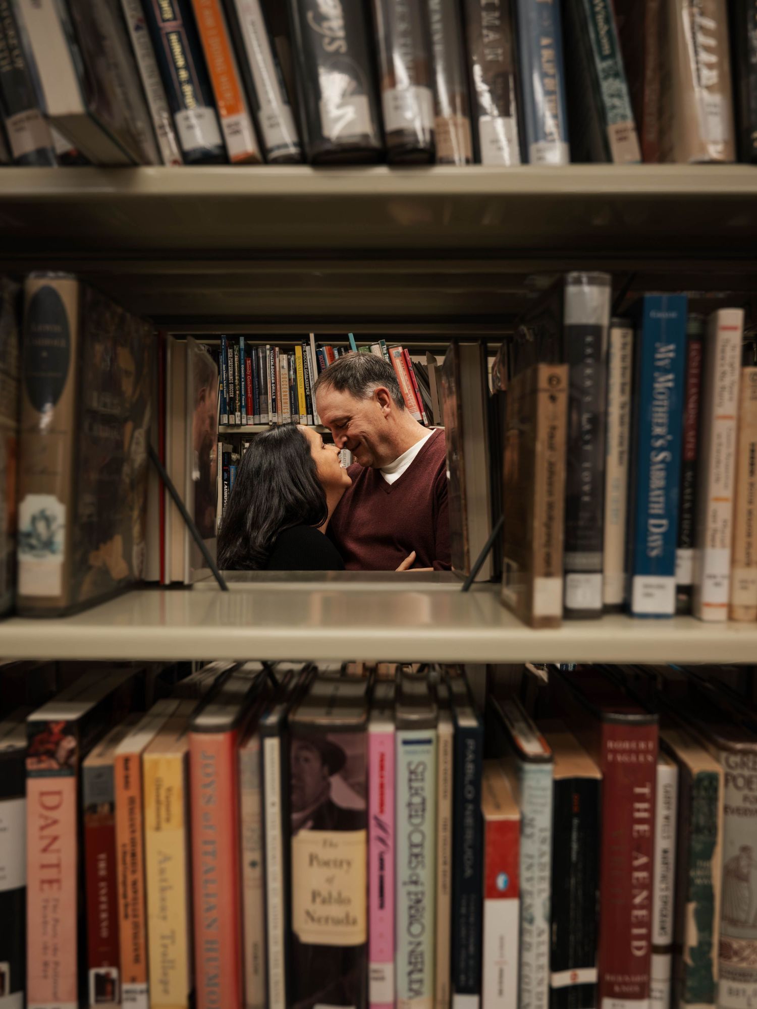 Romantic couple kissing seen through a gap in library bookshelves filled with colorful books.