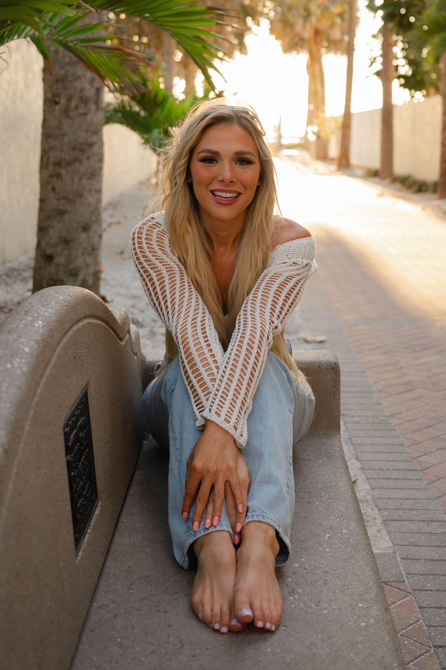Smiling blonde woman sitting on a bench along a palm tree-lined path at golden hour.