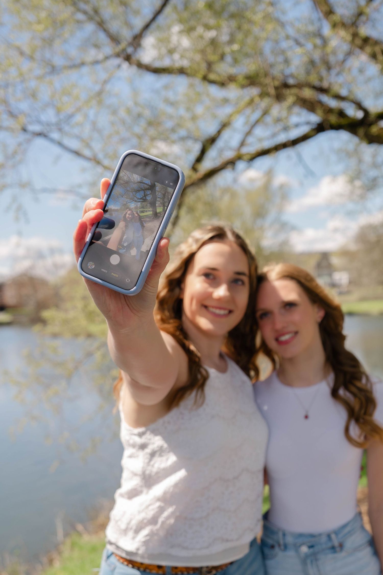 Two smiling young women take a selfie outdoors near a pond surrounded by spring trees.