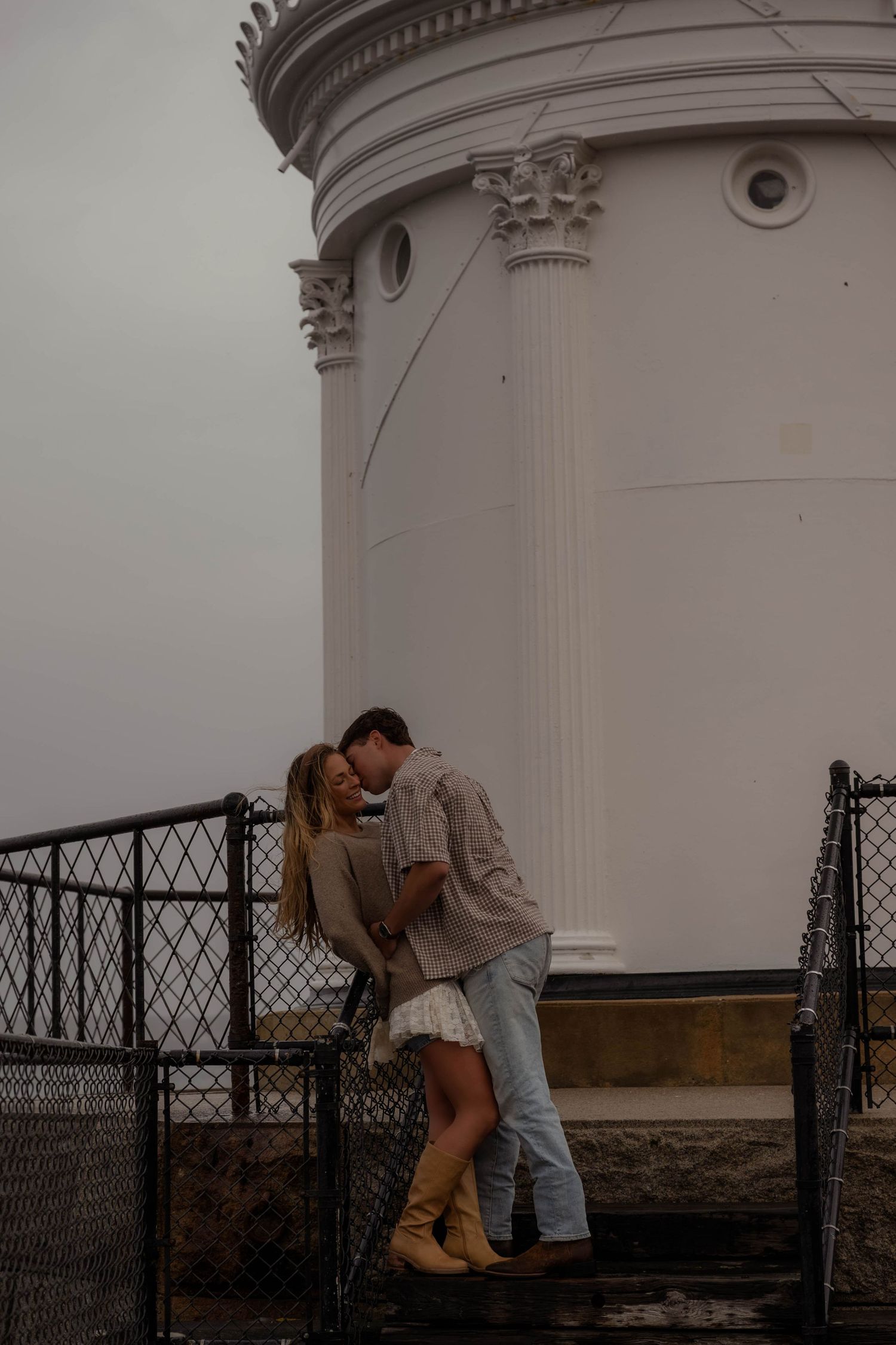 Romantic couple kissing near white lighthouse column on overcast day.