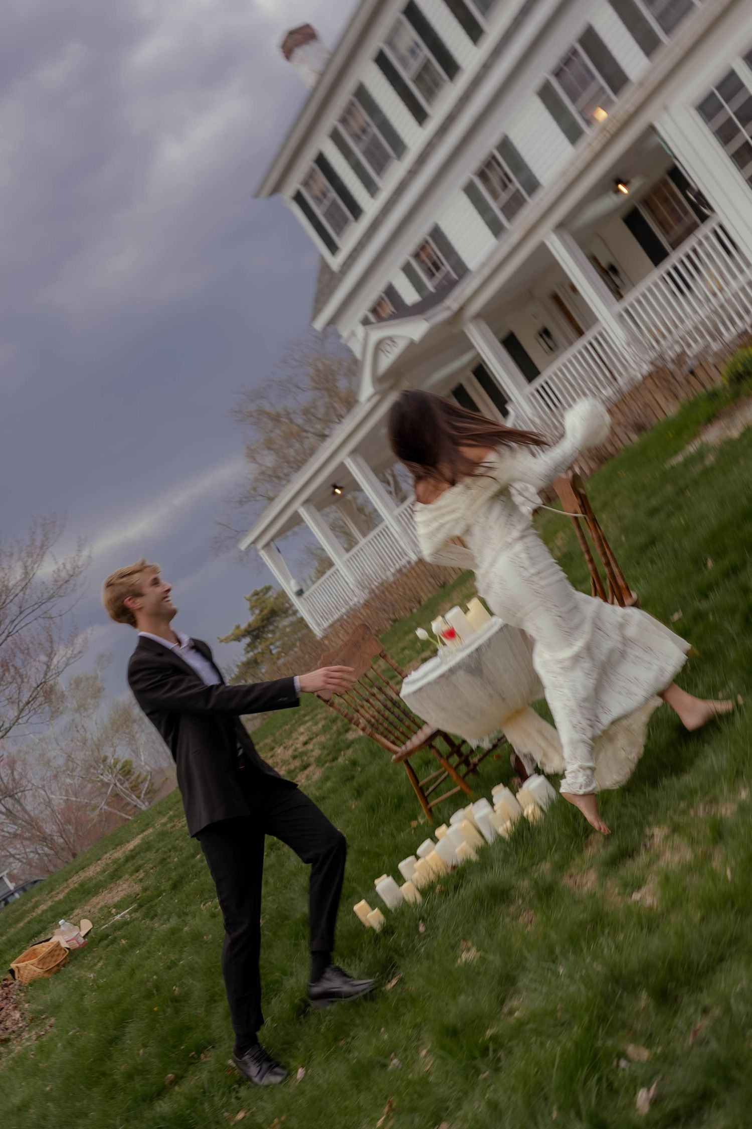 Bride in white dress and groom in black suit dancing joyfully on green lawn outside elegant white colonial house.