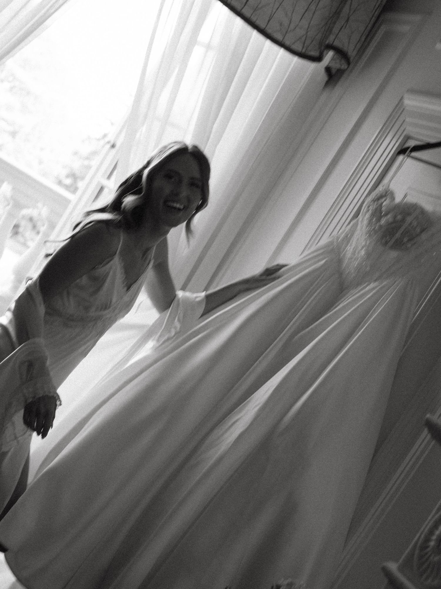 Black and white photo of joyful bride laughing beside her wedding dress hanging near bright window.