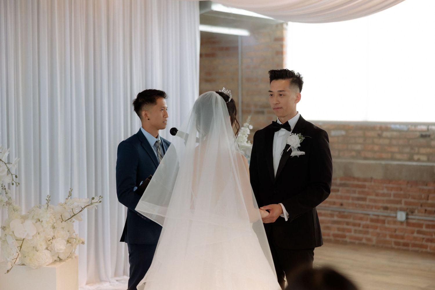 Groom in black tuxedo exchanges vows with veiled bride at a modern wedding ceremony with white draping.