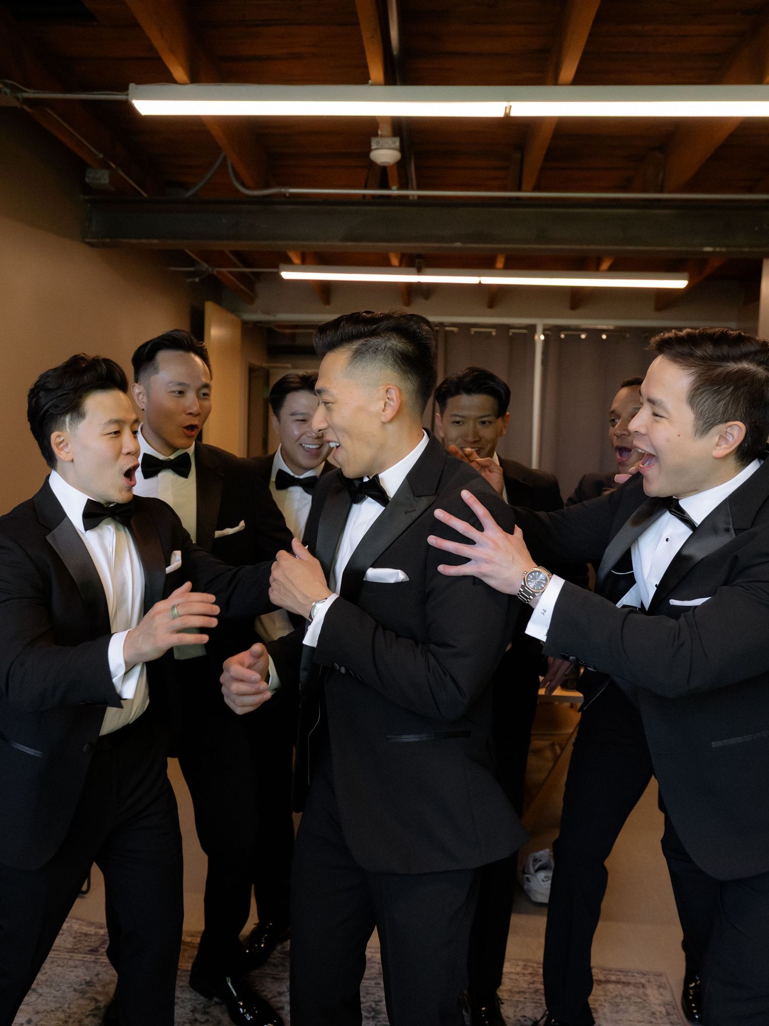 Groom surrounded by groomsmen in black tuxedos laughing together before wedding ceremony.