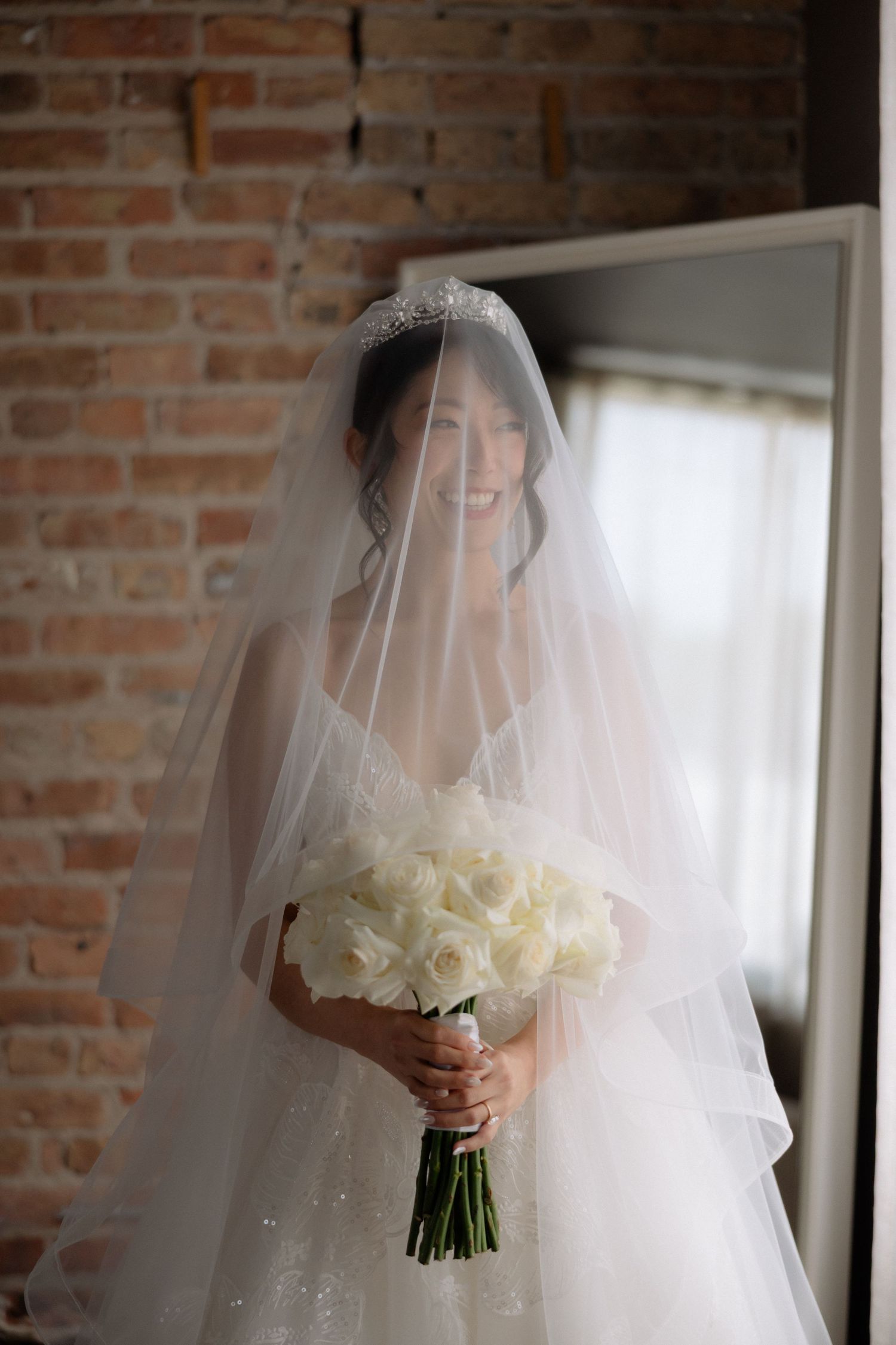 Smiling bride in white ball gown and veil holds white rose bouquet against exposed brick wall.