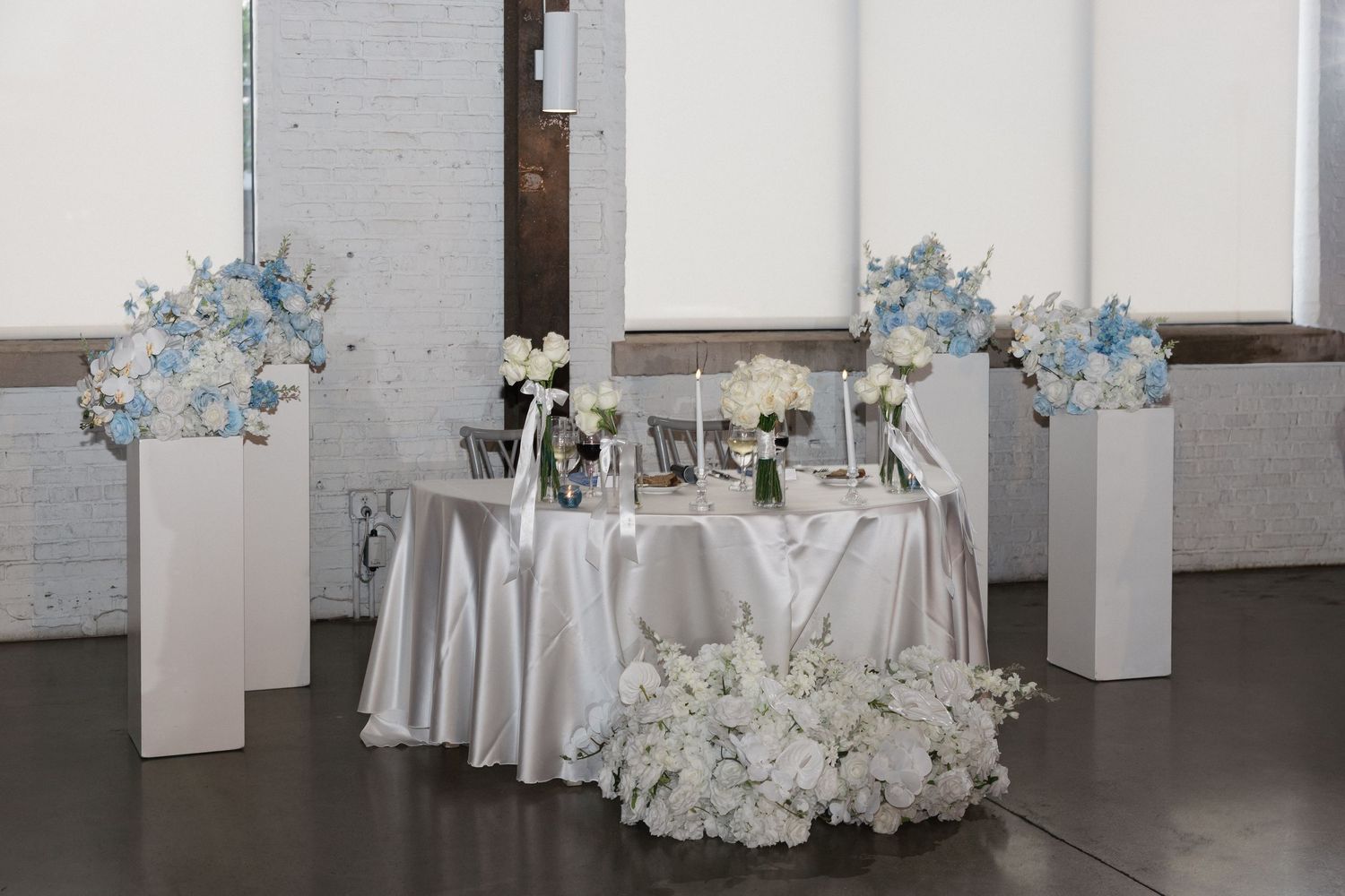 Elegant wedding sweetheart table with silver linen, white and blue floral arrangements on white pedestals in a bright venue.