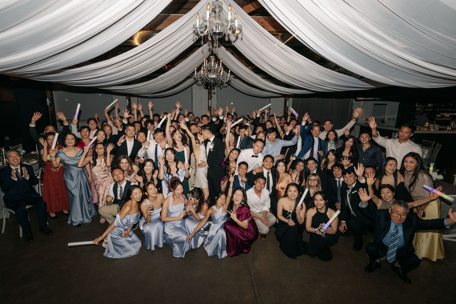 Large group of wedding guests posing together under draped ceiling at elegant indoor reception venue.