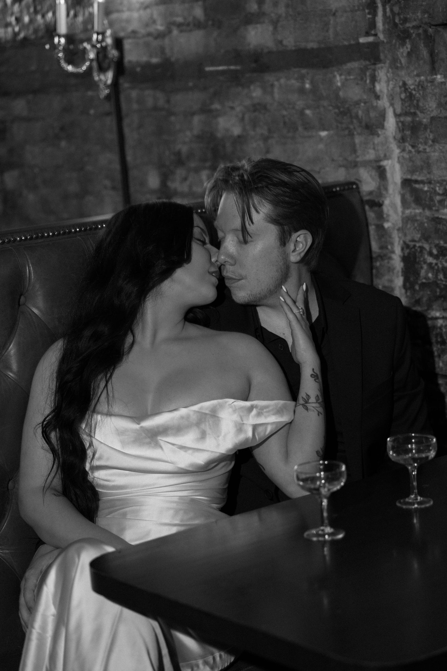 Black and white photo of a bride and groom sharing an intimate moment, seated at a table with wine glasses nearby.
