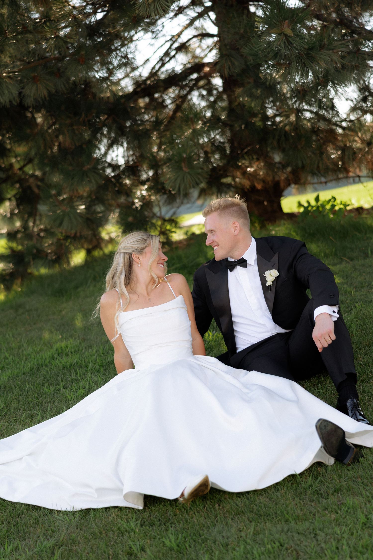 Bride and groom sitting on green grass under pine tree, smiling at each other, wearing wedding dress and tuxedo.