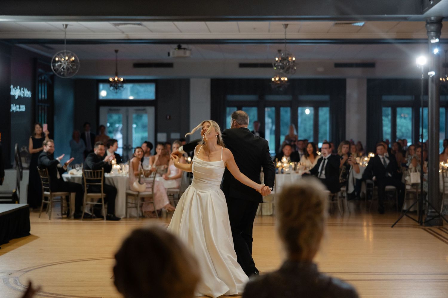 Bride and groom share their first dance on a wooden dance floor surrounded by seated wedding guests in an elegant venue.
