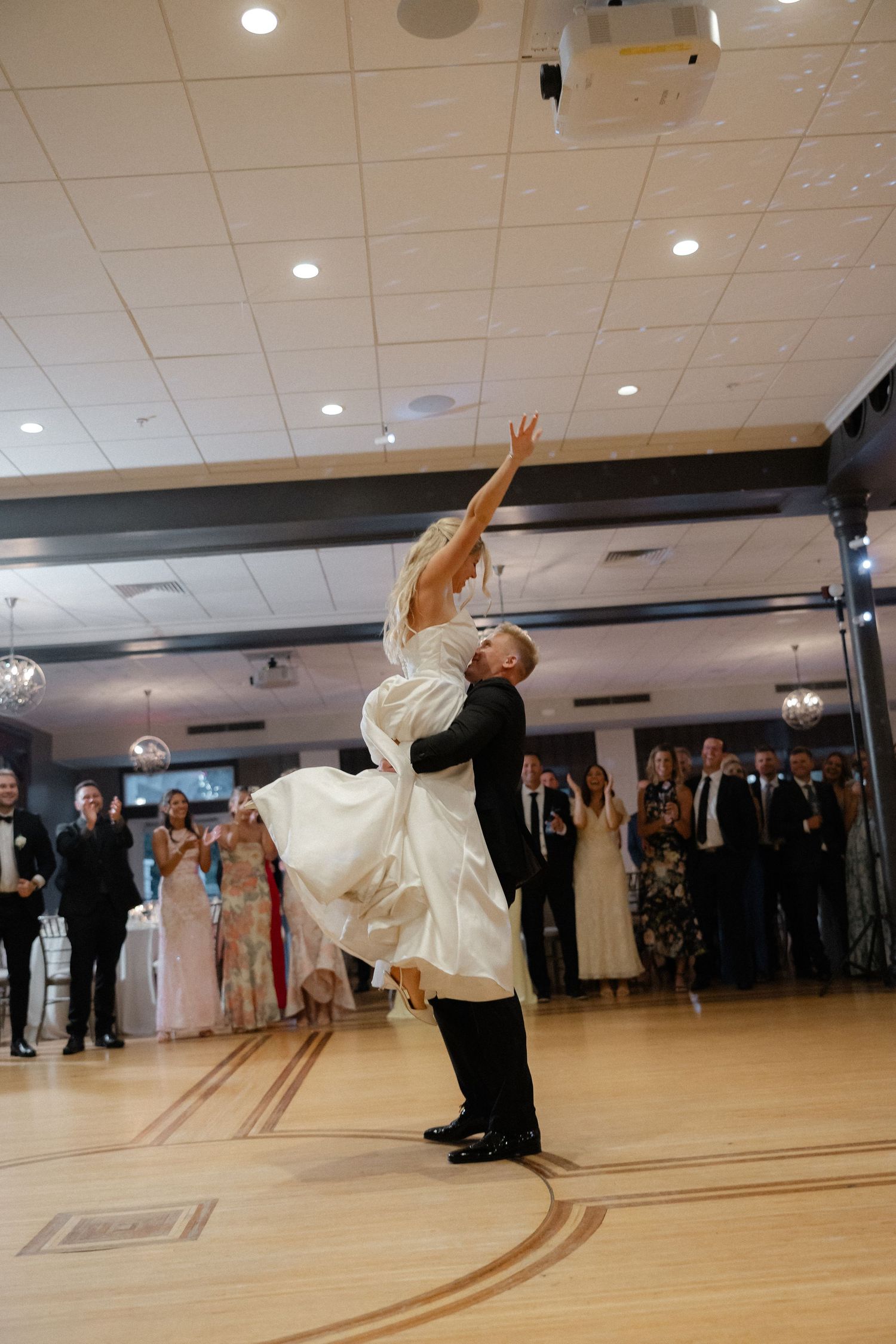 Groom lifting bride in white wedding dress during stunning first dance lift in elegant ballroom.