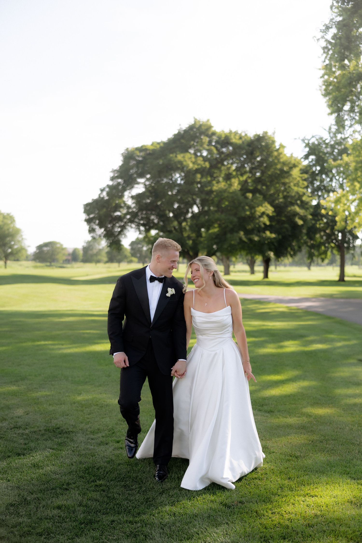 Bride and groom walk hand-in-hand across lush green lawn, smiling at each other in elegant wedding attire.